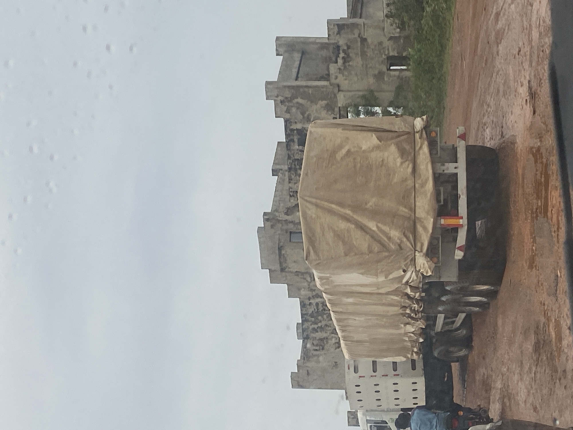 A convoy of vehicles, including a white truck with ventilation holes, a large flatbed truck covered with a tan tarp, and a motorcycle with a rider, is on a dirt road in Keffi, Nigeria. The road is muddy and appears to have puddles. In the background, a weathered, fortress-like building with several levels and irregular battlements stands under a cloudy sky. The building is made of concrete and shows signs of age and disrepair. The overall atmosphere is overcast and somewhat desolate.