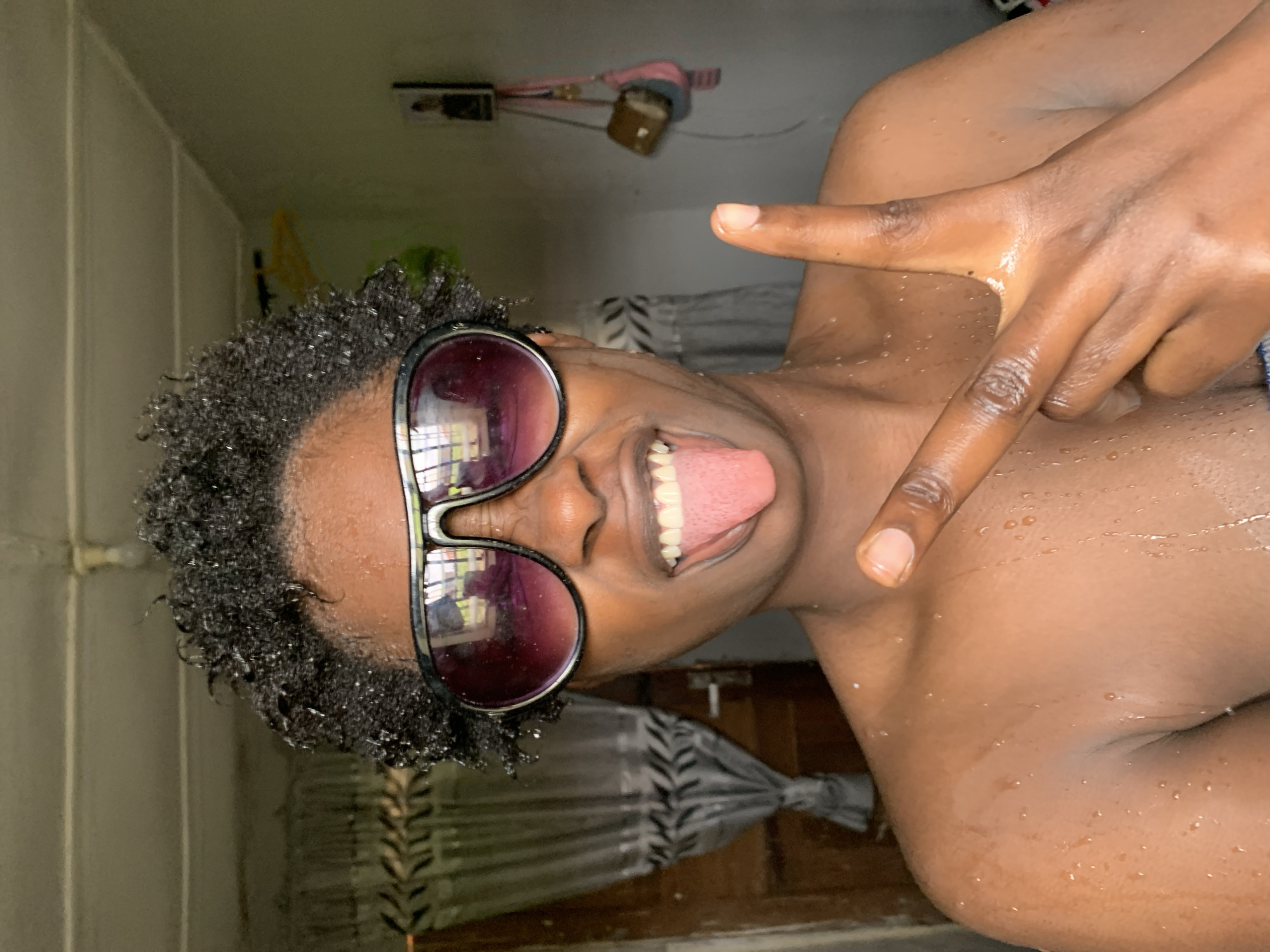 The media file is a close-up, vertically oriented, slightly tilted photograph of a person with dark skin, wearing sunglasses, and sticking out their tongue. The person is partially submerged in water, with water droplets visible on their skin and hair. Their hair is dark and curly, and the sunglasses have dark, reflective lenses. The person is making a gesture with their hand, forming a "Y" shape with their index and middle fingers. The background is out of focus but appears to be an indoor setting, possibly a bathroom, with a tiled wall and a shower curtain visible. There are some items hanging on the wall in the upper left corner. The lighting suggests it could be daytime, but it's difficult to determine precisely. The person's expression appears playful or mischievous. There is no visible text in the image. The location context is Rafin Par, Nigeria.
