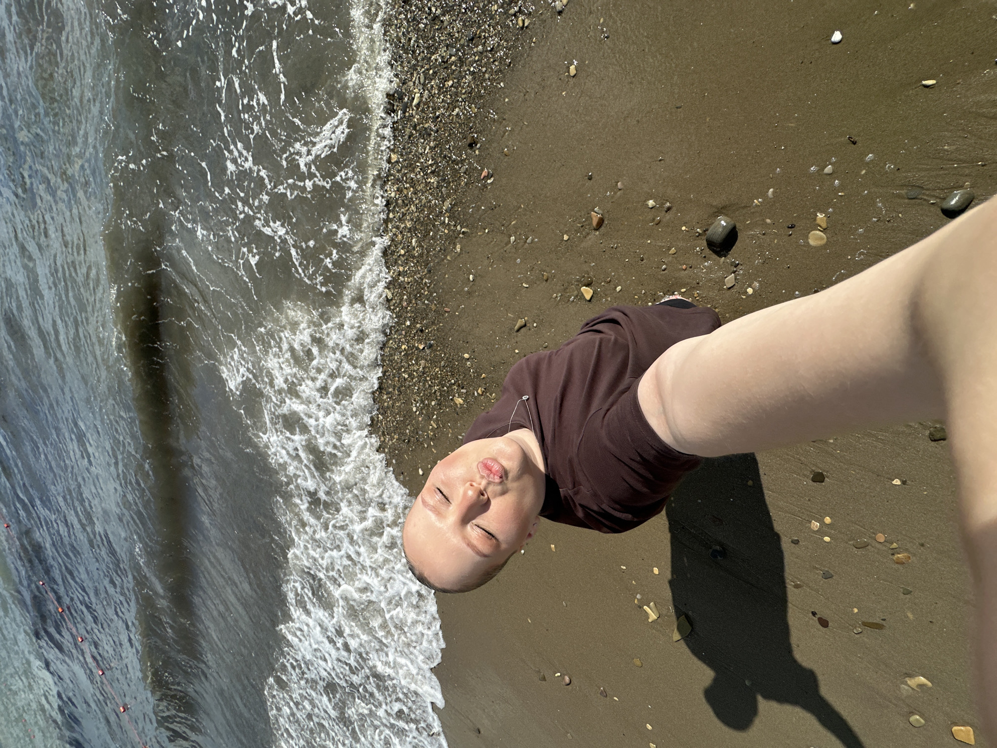 In Lermontovo, Russia, a young person with a shaved head, wearing a brown shirt, is taking a selfie on a beach. Their eyes are closed and lips are puckered as if for a kiss. The person's arm, holding the camera, is extended towards the viewer, obscuring part of their body. The background shows a rough, pebble-strewn beach meeting the dark, choppy water of the Black Sea, with small orange buoys visible on a rope in the distance. The sun casts a long shadow of the person onto the wet sand.