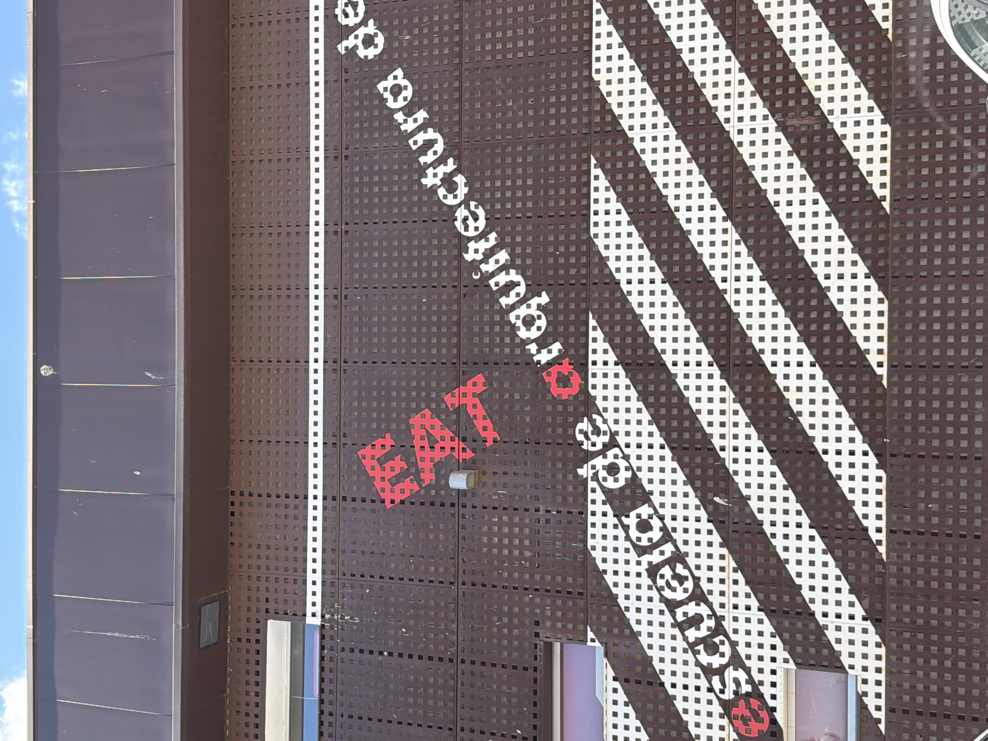 The image shows the exterior of a building with a prominent facade featuring a dark brown, perforated metal screen. Red and white text is applied to this screen. The red text, "ETS", is positioned on the upper left portion of the screen, oriented vertically. To the right and below, white text reads "escuela de arquitectura de". This white text is arranged diagonally across the screen, following the pattern of alternating dark and light stripes formed by the perforations in the screen. The building appears to be modern, with visible windows and what looks like a ventilation grate above the perforated screen. The sky above is blue with some clouds. The overall impression is one of an architectural or educational institution in Toledo, Spain, given the text.