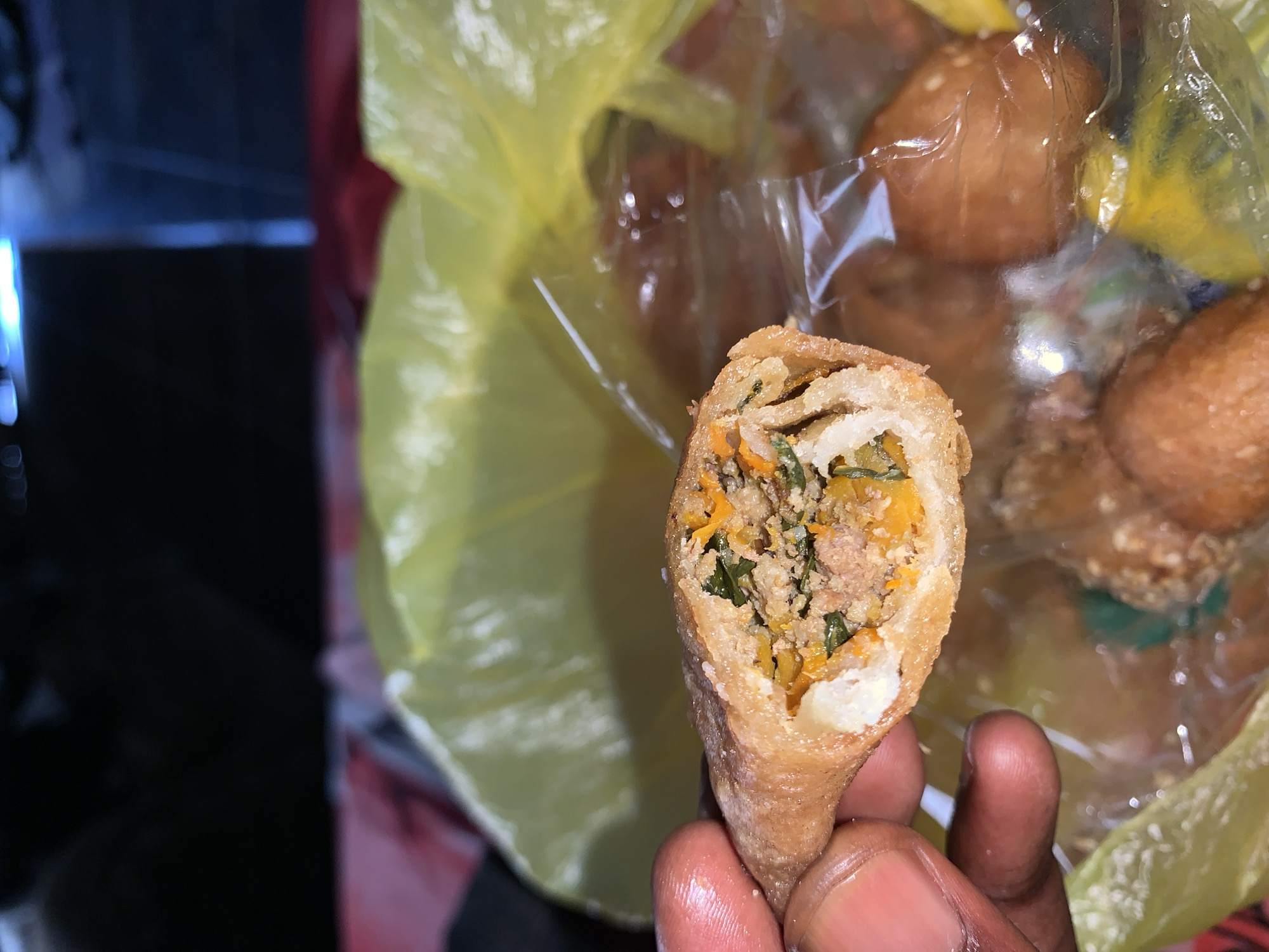 A close-up, eye-level, outdoor shot shows a person's dark-skinned hand holding a fried spring roll. The spring roll is cut in half, revealing its filling of ground meat, chopped green leafy vegetables, and julienned carrots. The spring roll is golden brown and appears to be crispy. In the background, out of focus, are more spring rolls also in a yellow plastic bag. The scene is likely in Jos, Nigeria, a city known for its street food culture.