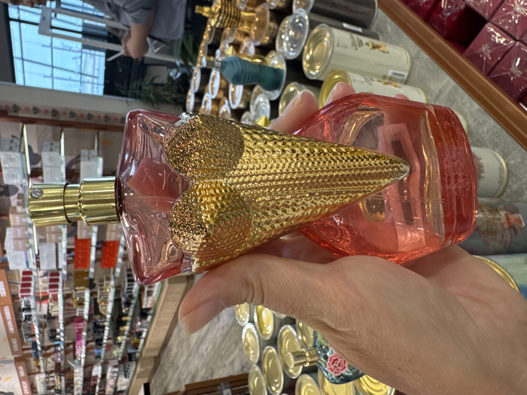 A hand holds a pink perfume bottle with a gold, patterned top. The background includes various products on shelves and other perfume bottles on a counter. The scene appears to be inside a shop selling perfumes and cosmetics. Based on the visible products on the shelves, it could be a location that features a variety of brands of cosmetic and personal care products. Without additional cues, specific locations in Tashkent, Uzbekistan, cannot be ascertained, but it is likely inside a commercial retail store.