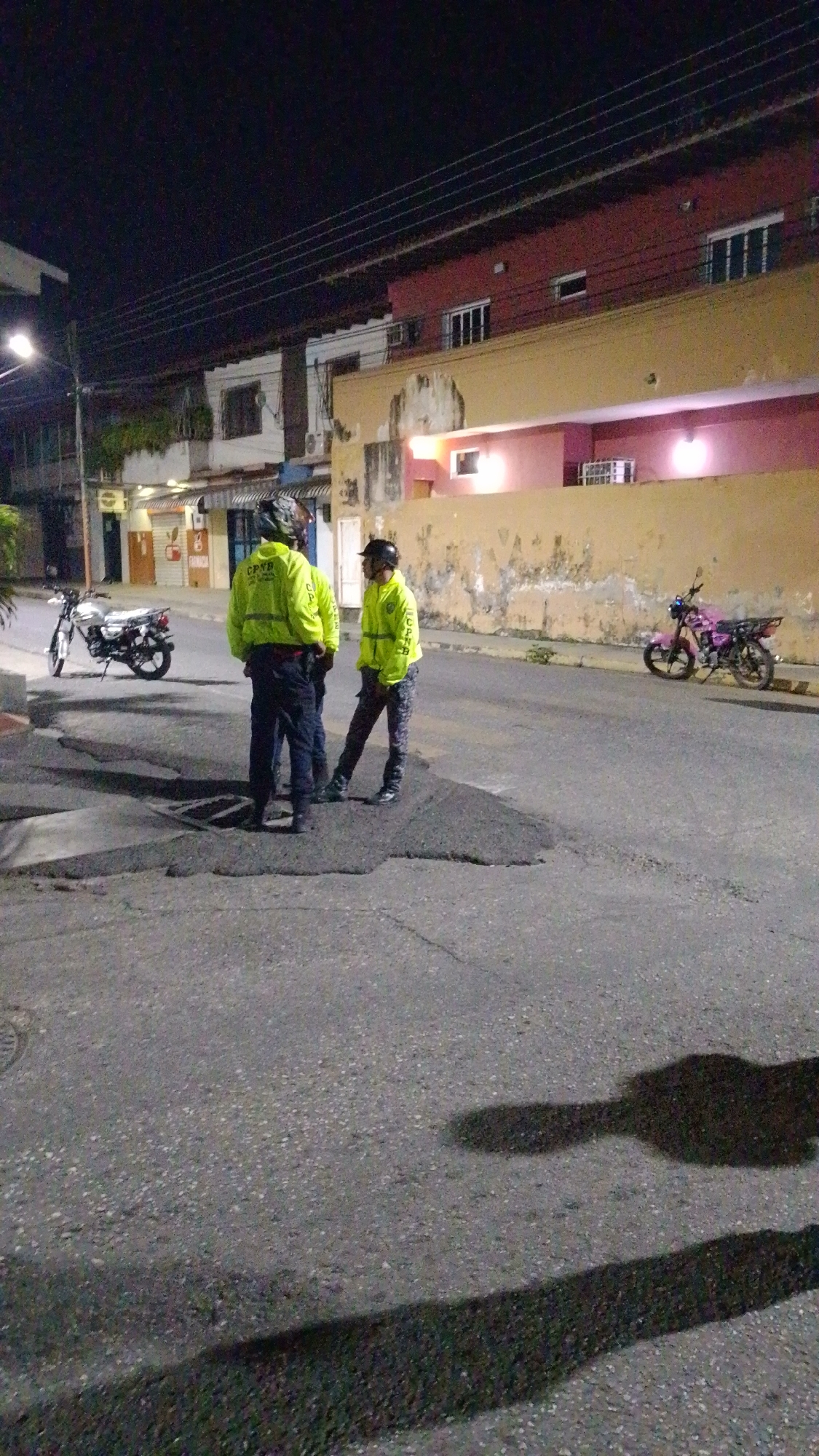 This nighttime image depicts an urban street scene in Tinaquillo, Venezuela, featuring three individuals, two motorcycles, and buildings. The three individuals, seen from behind, are wearing bright yellow-green jackets with dark pants and helmets. Text visible on their jackets includes "CPNP DIV. REGENERACIÓN" and "CPNB," indicating they are members of the Bolivarian National Police Corps (Cuerpo de Policía Nacional Bolivariana). They are standing and conversing in the middle of the asphalt street, which shows patches and an open manhole cover nearby.
On the left side of the street, a silver motorcycle is parked. Further down on the right, a pink motorcycle is also parked along the curb. The background consists of various multi-story buildings with different facades, including a prominent red building, a yellow-orange wall, and white structures. Some buildings have windows, and visible awnings suggest commercial establishments. The sky is dark, indicating night, with artificial lights from streetlights and building interiors illuminating parts of the scene and casting shadows on the street. The overall impression is a quiet, routine evening on a Venezuelan city street.