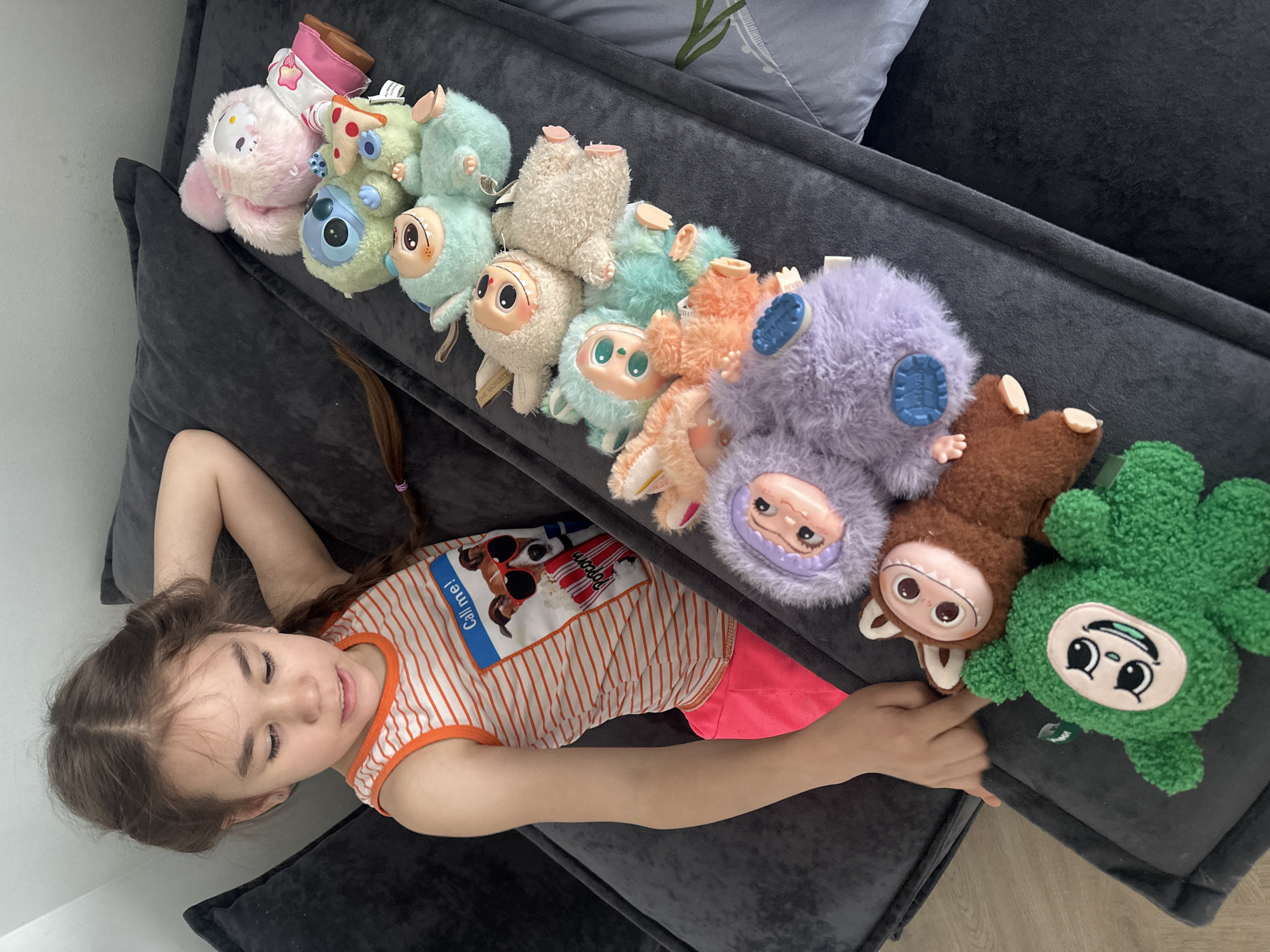 A young girl with light skin and brown hair, styled in two braids, lies on a dark gray upholstered sofa. She is wearing a striped orange and white tank top and bright pink shorts. To her right, arranged in a line along the sofa back, are a collection of approximately twelve small, plush toys of various colors and designs. The sofa is situated indoors, possibly in a residential setting. The location in the city of Kharnavy, Russia is not discernible from the image.