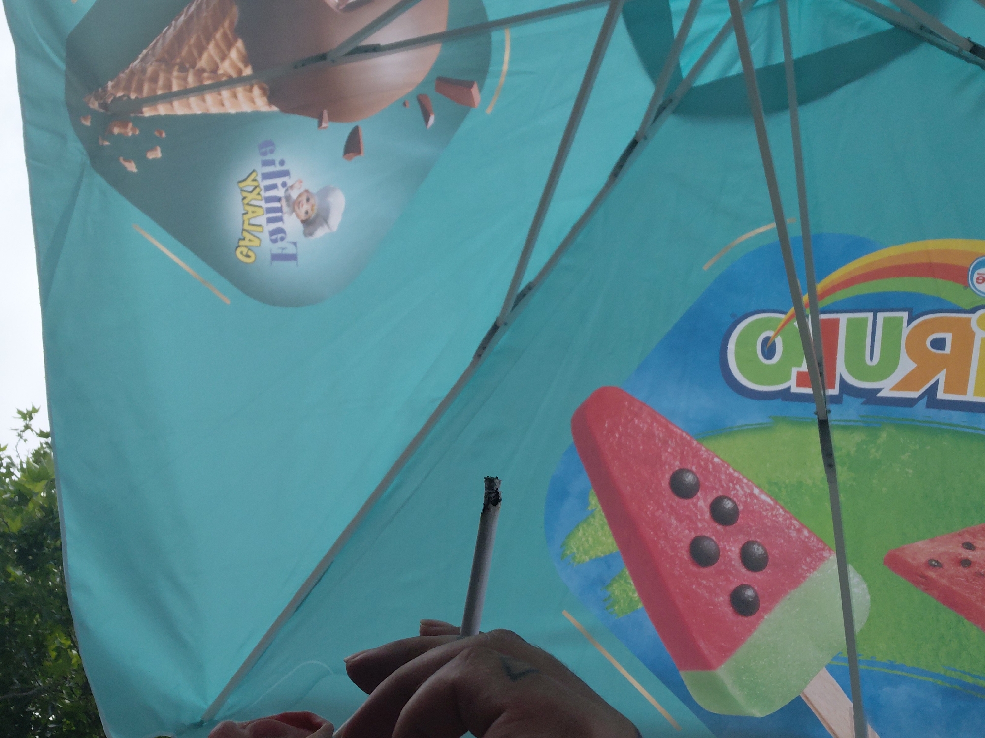 The scene depicts a view from underneath a teal-colored umbrella in Kostievo, Bulgaria. The umbrella's underside features advertisements for ice cream. On the upper right section, the partially visible "BUBBI" logo with rainbow colors is present, alongside an illustration of a watermelon-slice-shaped popsicle with black "seeds" on a green base, on a stick. Another red triangular ice cream form is visible above it. On the lower left section, an image of a chocolate-dipped ice cream cone with a waffle cone is displayed. Adjacent to the cone is a reversed text logo, "silimeT YXAJAS" (likely "Familia GALAXY"), accompanied by a cartoon chef character. To the right, an adult hand is holding a lit cigarette, with visible ash at its glowing tip. The metal spokes and structure of the umbrella are visible, crisscrossing the fabric. Below the umbrella, a bright, clear sky is visible, with green foliage and tree branches appearing along the bottom edge, indicating an outdoor setting.