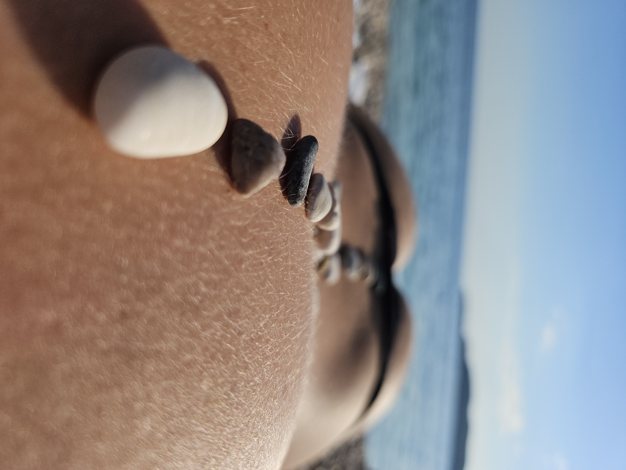 A close-up, slightly tilted image shows small stones placed on human skin, with a beach and water visible in the blurred background. The skin appears tanned and has fine hairs. Several small, smooth stones of varying colors, including white and gray, are arranged in a line. The background shows a blurred coastline and blue sky. The location appears to be a beach, possibly in an area near Unknown, Unknown.