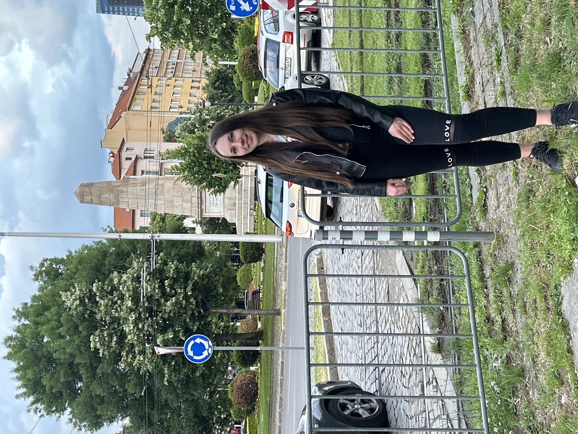 The image displays a young woman posing outdoors on a street in Sofia, Bulgaria, during daytime under a partly cloudy sky. She has long, dark hair and is smiling at the camera, standing beside a grey metal fence. She wears a black leather jacket over a white top, black leggings with the word "LOVE" printed on them, and black sneakers.
Behind the woman and the fence, a cobblestone-patterned walkway leads to a street. A white car with the Bulgarian license plate "CB 2756 KT" is visible further down the road. In the mid-ground to the left, a tall, grey stone monument or obelisk stands amidst trees and greenery. The street is lined with multi-story buildings, featuring facades in shades of yellow, orange, and white, characteristic of European architecture. A modern glass skyscraper is visible in the distant background. Two blue circular road signs, indicating a mandatory roundabout or circular traffic, are present along the street. The scene depicts a casual moment in a well-maintained urban setting.
