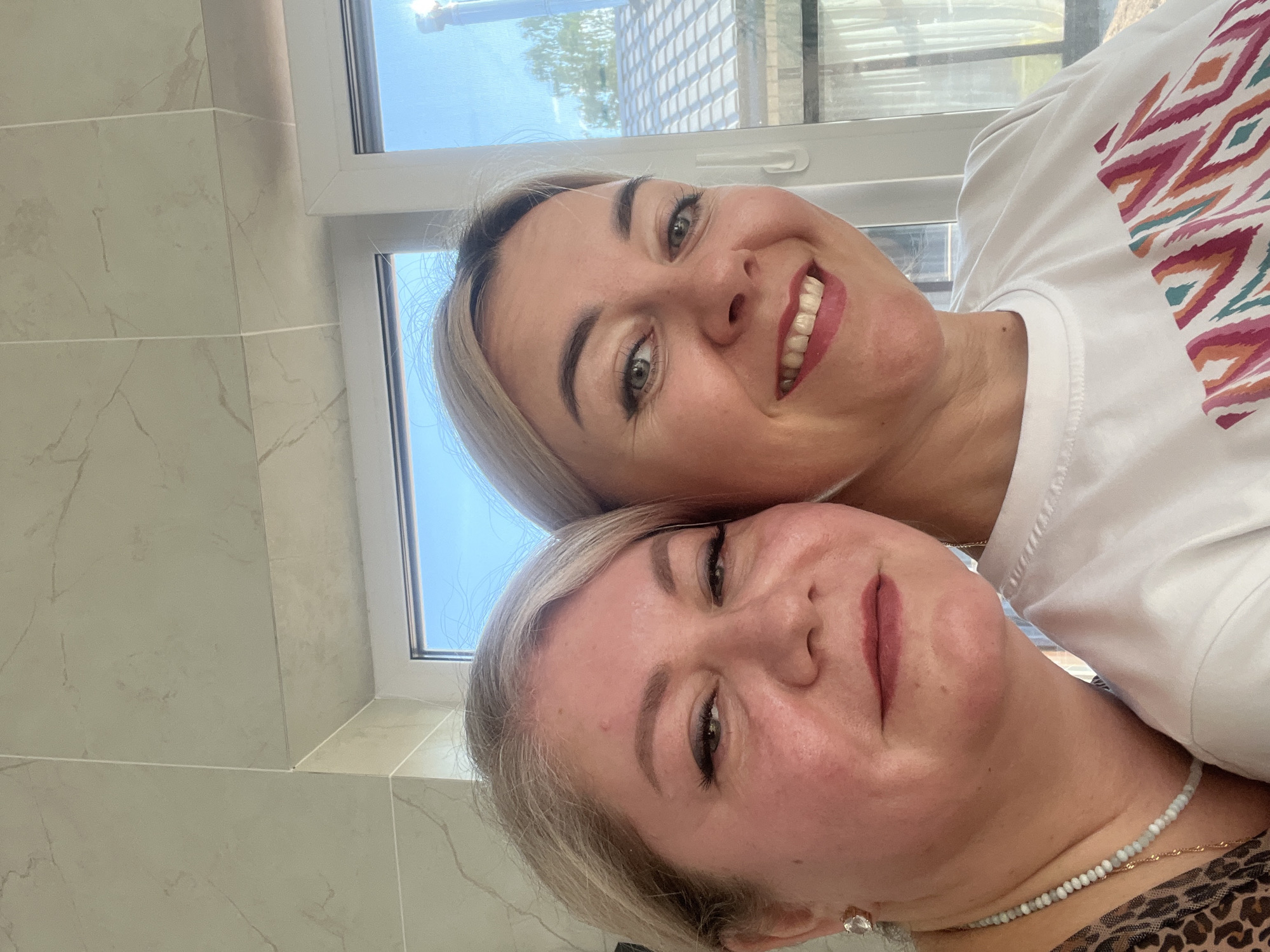 Two women are posing for a selfie indoors, near a window. The woman on top has blonde hair and a white t-shirt with a colorful geometric pattern. She is smiling broadly, exposing her teeth. The woman below has blonde hair, and a beaded necklace. She is smiling gently. The background shows a tiled wall and a window looking out to a view that appears to be in Novaya Chernushka, Russia.