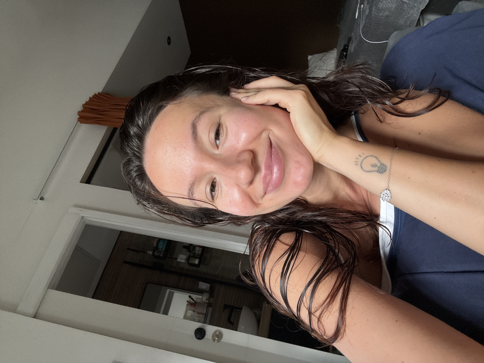 A young woman with wet, dark hair and bare shoulders smiles gently, resting her chin on her right hand. She is wearing a dark blue tank top. A tattoo of a light bulb is visible on her left forearm, accompanied by a silver bracelet. The background appears to be an indoor domestic setting, possibly a bedroom, with a glimpse of a bathroom through an open doorway on the left. The lighting is soft and natural. There is no indication of the image being located in Tel-Aviv, Israel.