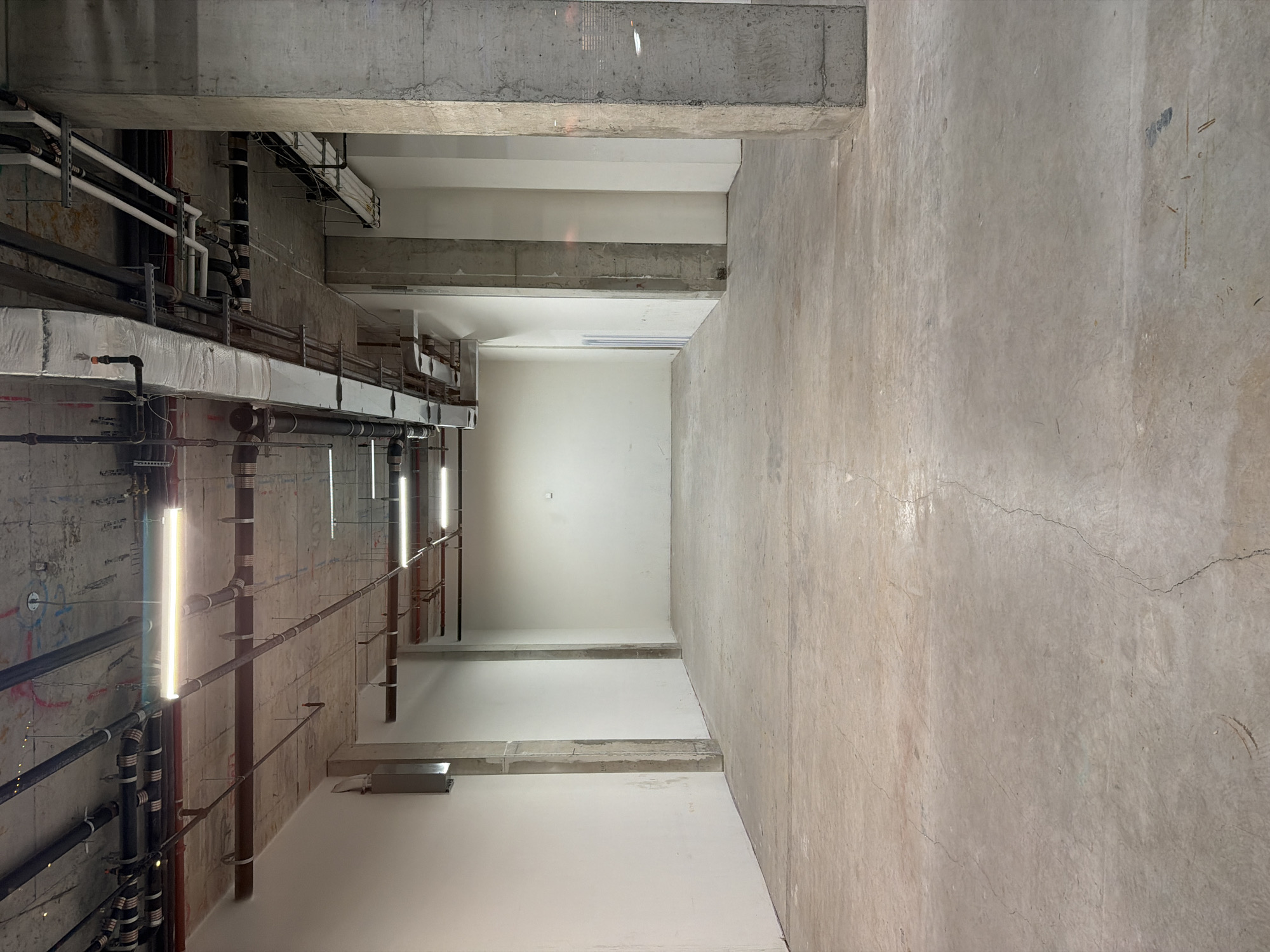 The image depicts an unfinished interior space. The left side features exposed pipes, ducts, and electrical conduits against a concrete wall with fluorescent tube lights. The right side showcases a plain concrete wall with cracks. The space appears to be a long, narrow corridor or room, possibly in a commercial or industrial building. There are no people visible. The location is unknown.