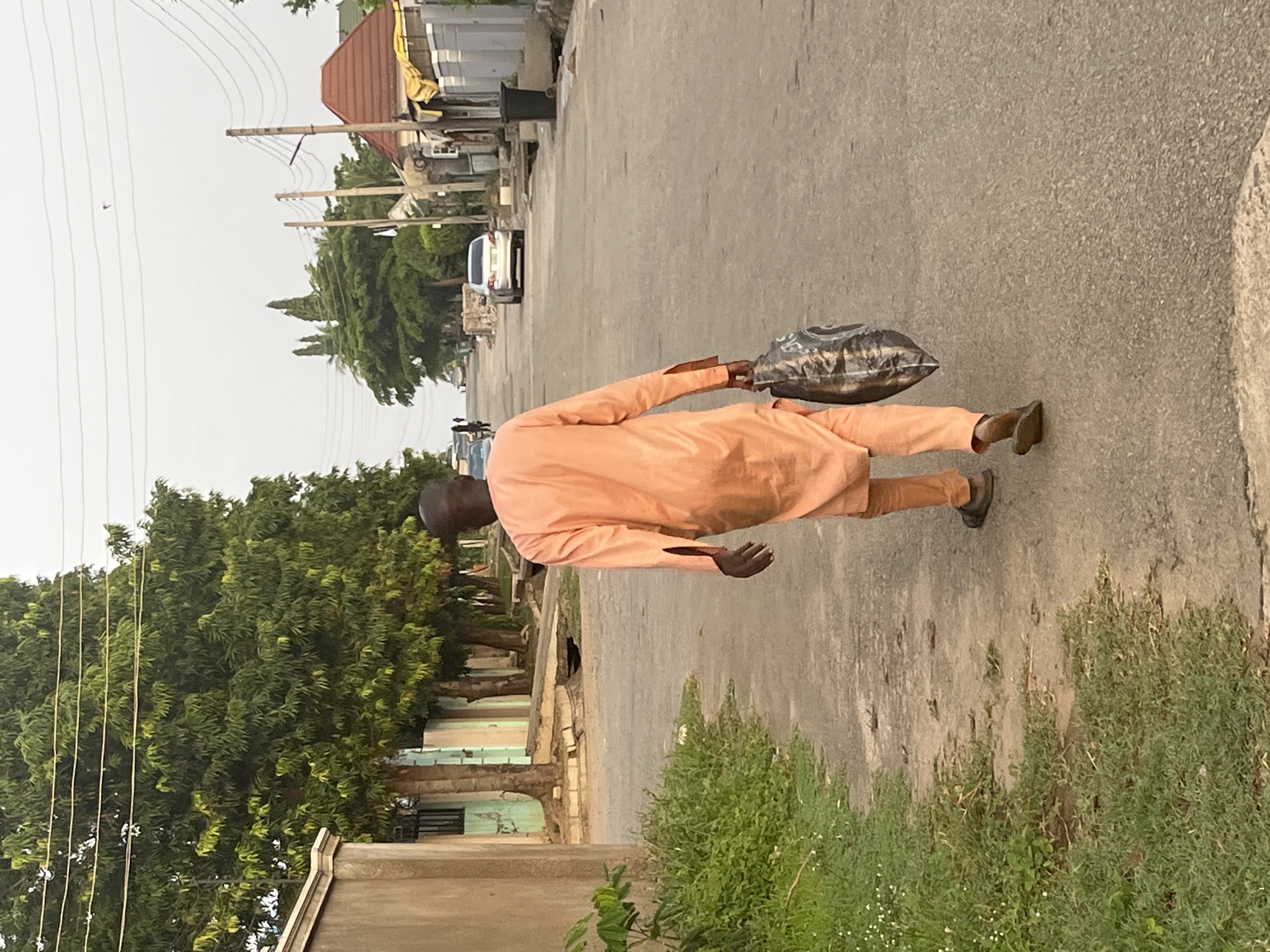 A man wearing an orange traditional garment walks on a paved road in Abuja, Nigeria. He carries a crumpled, patterned bag in his left hand. The road is lined with trees and residential buildings. In the background, cars and electrical wires are visible.