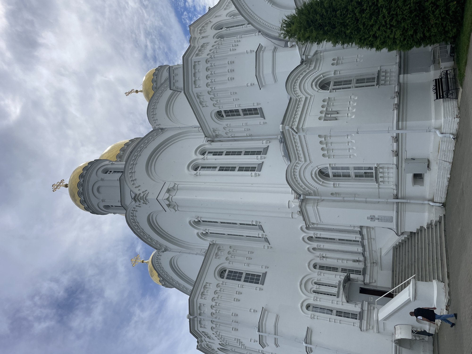 The image shows a low-angle view of a large, white, Orthodox church with golden domes, located in Diveyevo, Russia. The church has intricate architectural details, including numerous arched windows and decorative carvings. The sky is partly cloudy, with patches of blue visible. In the foreground, a man in a dark vest and blue jeans is walking down a wide set of stairs leading to an entrance of the church. To the right of the stairs, a tall, green coniferous tree stands prominently. A black bench and a silver trolley are visible near the base of the church. The ground is paved with grey stone. The scene appears to be during daylight.