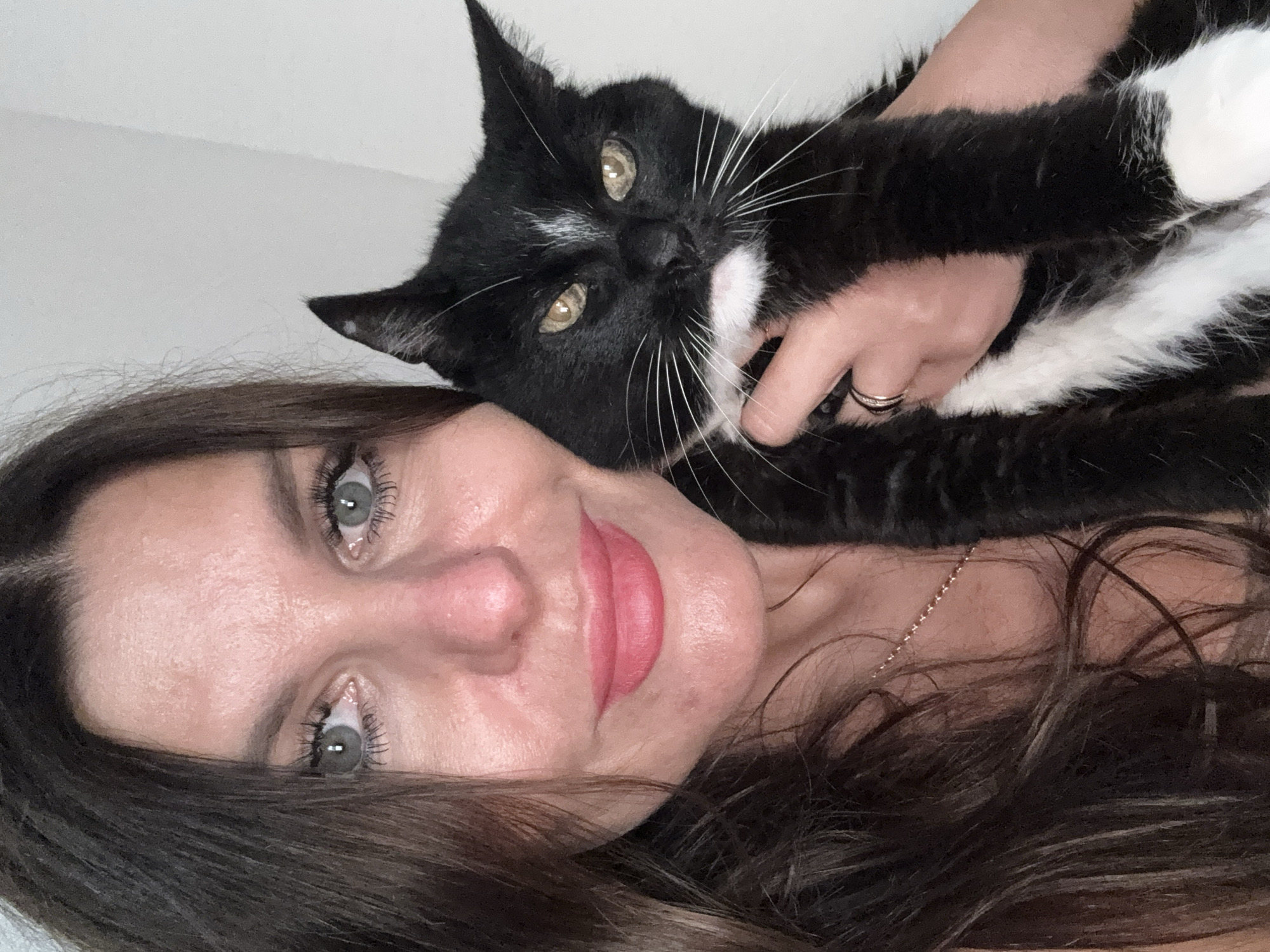 A woman with long, dark brown hair and blue eyes is holding a black and white cat. The woman is wearing a light-colored top and a gold necklace. The cat has green eyes and white whiskers. The background is a plain white wall. The image appears to be a selfie taken indoors.