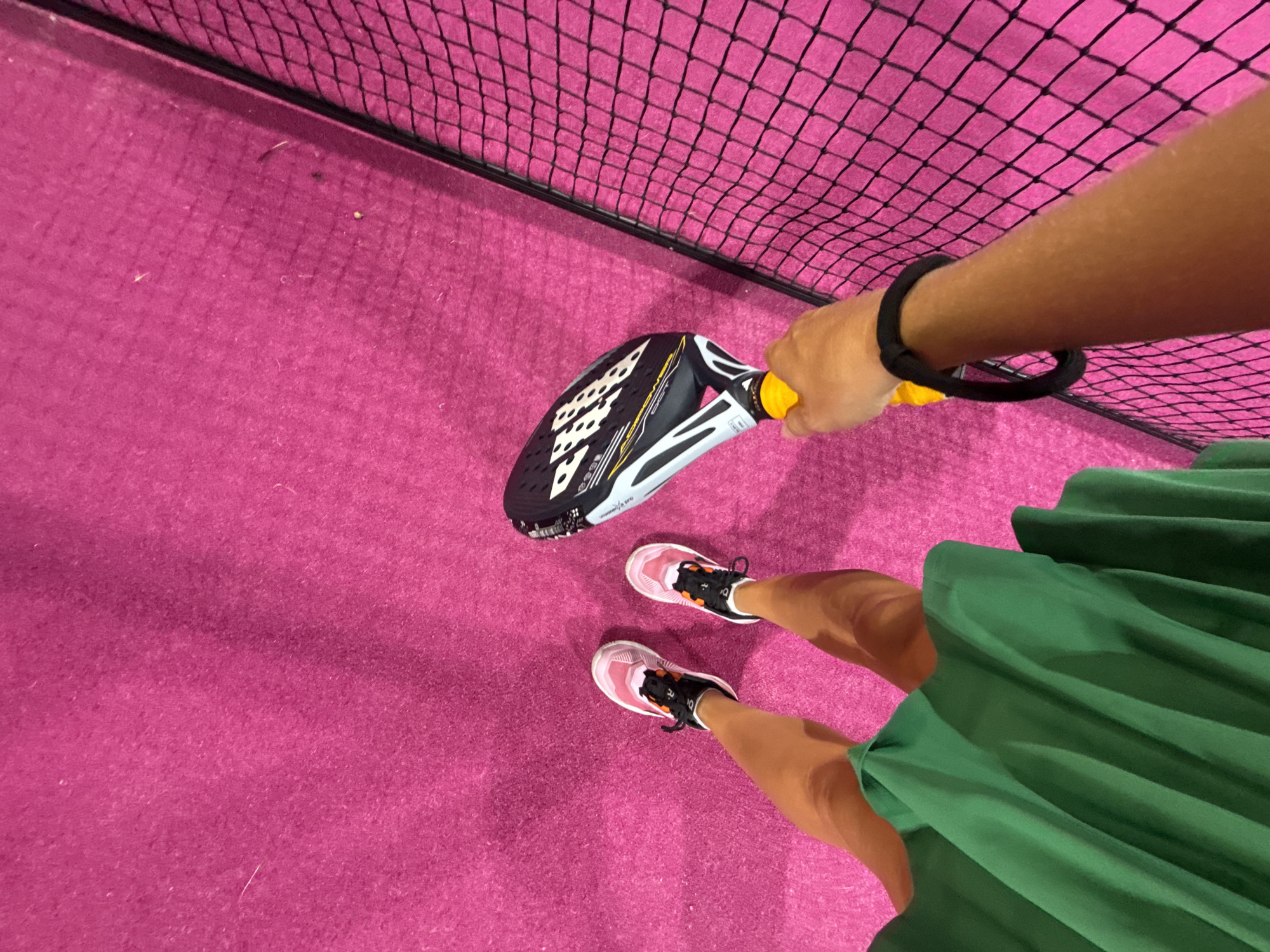 An overhead, high-angle, first-person perspective shot shows a person standing on a bright pink surface, possibly a sports court with a net on the right. The person's legs are visible, clad in athletic shoes with pink and white accents, and a flowing green skirt. The right hand is extended forward, holding a black and white paddle with yellow accents. The background is a solid pink surface. There is no indication of the location being in the city of Ko Samui District, Thailand.