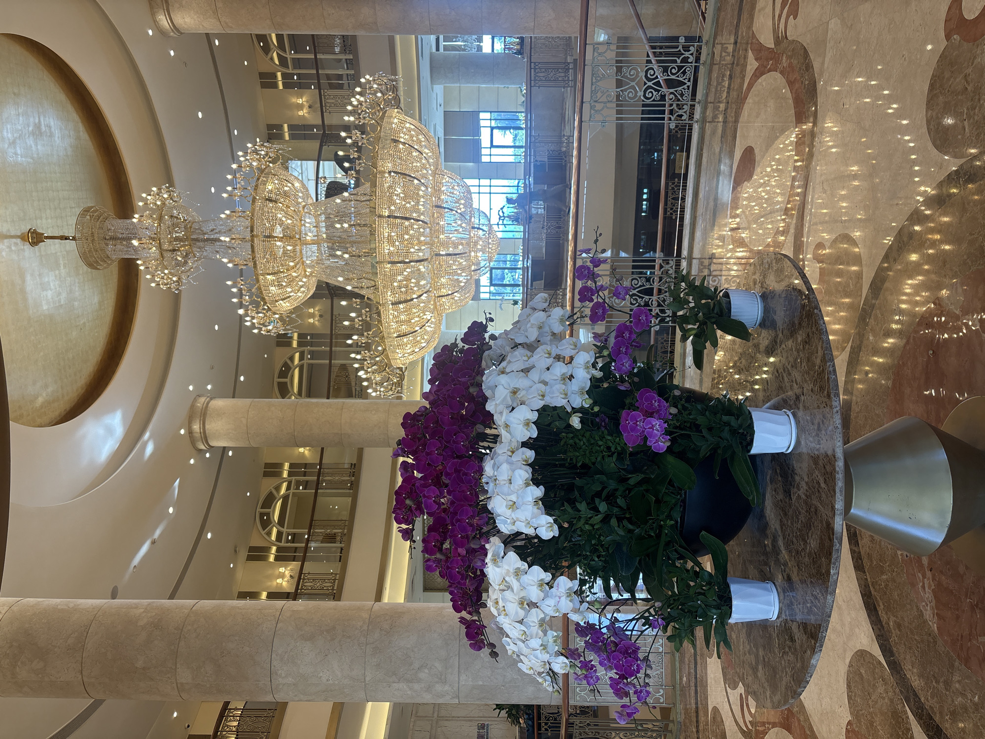 This image depicts the opulent lobby of a hotel in Da Nang, Vietnam. A massive, multi-tiered crystal chandelier hangs from a gold-accented domed ceiling, illuminating the space. Large marble columns support the upper levels, where balconies with ornate railings overlook the lobby. In the foreground, a round marble table displays a grand arrangement of white and purple orchids in a large black planter, flanked by two smaller white potted orchids. The polished marble floor features intricate inlays that reflect the ambient light and the chandelier's glow. The background reveals an upper floor with windows offering glimpses of the exterior and a reception area. A lone figure can be seen walking in the distance on the upper level.