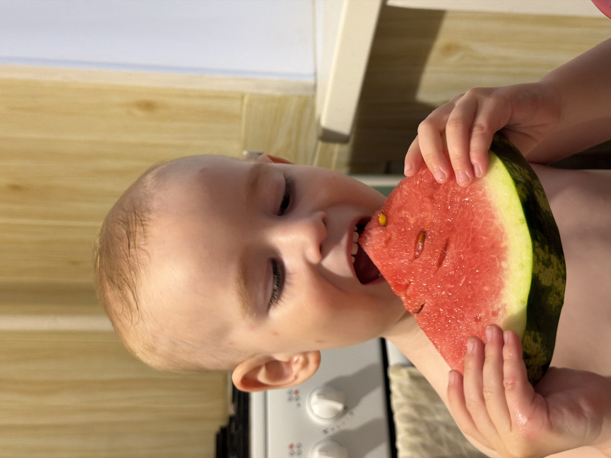 A young child is holding and eating a slice of watermelon. The child appears to be indoors, with a stove and kitchen cabinets visible in the background. The location is Chaikovsky, Russia. The child is fair-skinned with light hair and is holding the watermelon slice with both hands, taking a bite. The watermelon is red and green with several seeds. The child's face is expressive, indicating enjoyment of the watermelon. The environment seems to be a domestic kitchen setting.