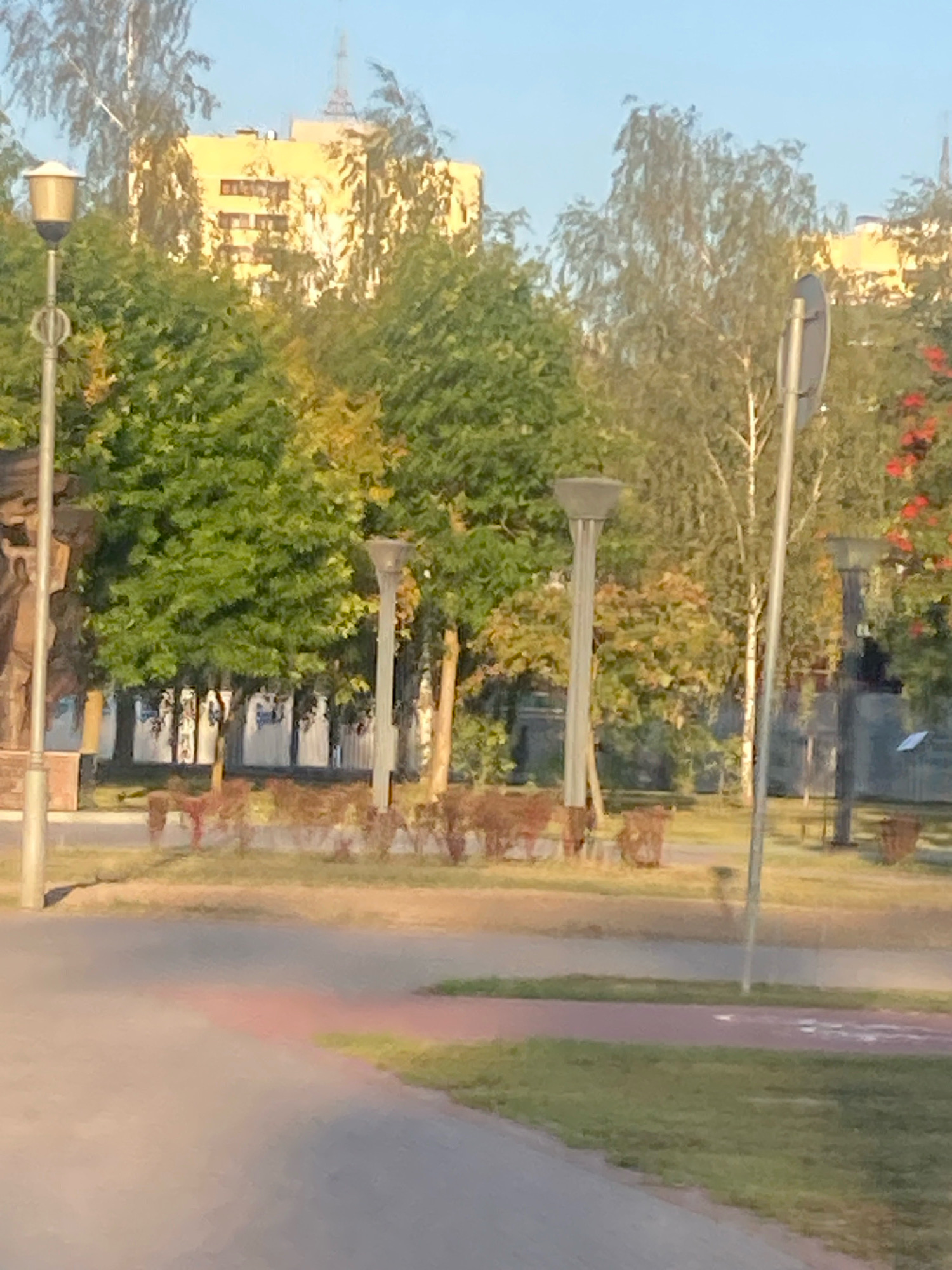The image shows a park area in the city of Brest, Belarus, bathed in natural sunlight. In the foreground, a paved path curves through a grassy area. In the middle ground, several lampposts with modern, geometric shades are visible, interspersed with trees and decorative bushes. A traffic signpost stands to the right of center. In the background, a tall, yellow, multi-story building with visible windows is partially obscured by lush green trees. The sky above is clear and blue. The overall impression is one of a well-maintained urban park.