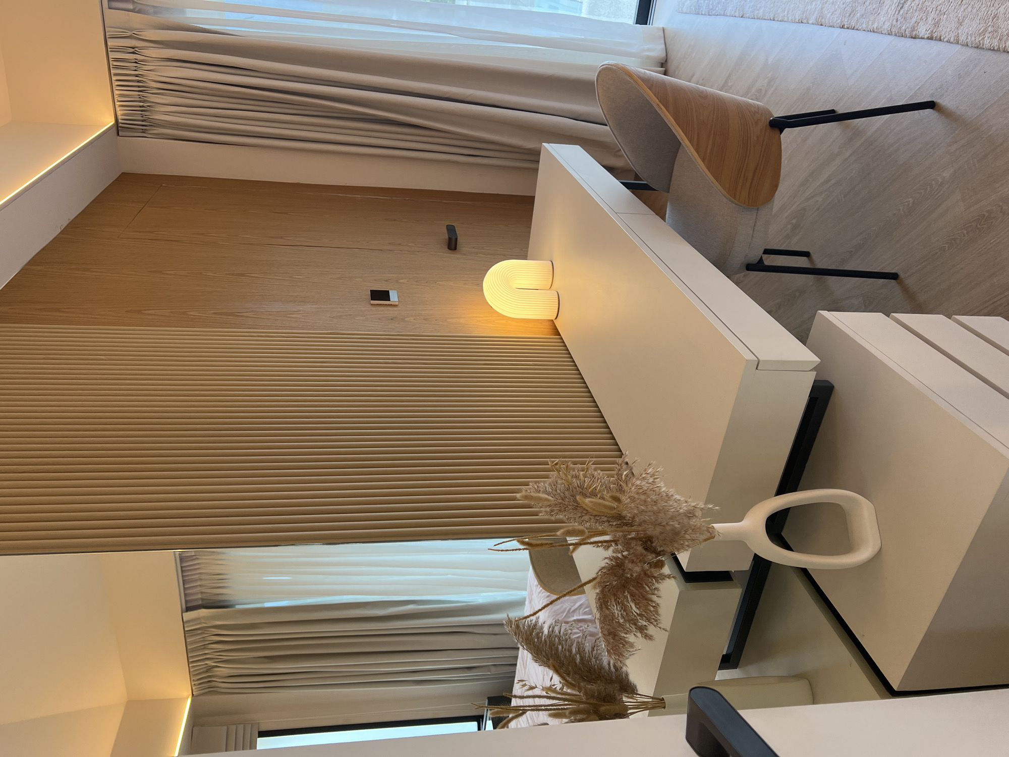 The image shows an interior, likely in Baan Bang Rak, Thailand, given the implied location.
**Environment:** A modern interior with a beige/light wood-paneled wall with vertical grooves. Light-colored curtains partially obscure a window. A white surface, possibly a desk or counter, extends into the room. The floor appears to be light wood.
**Objects:** A bent plywood chair with black metal legs is visible. A curved, ribbed light fixture is mounted on the wall. A vase with dried floral arrangements is placed on a white surface in the foreground. A mirror reflects a bed and window, suggesting a bedroom.
**Overall:** The image captures a scene with modern, minimalist design elements, incorporating natural wood tones and white surfaces. The reflected image of the bed and window indicates that this interior is likely a bedroom.