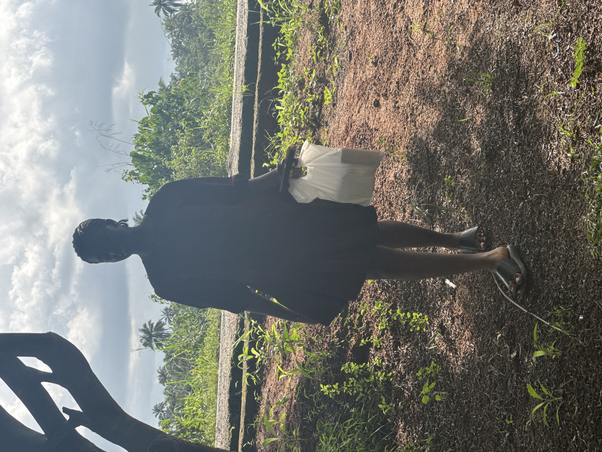 A person stands in a natural outdoor setting in Ihiagwa, Nigeria. The ground is covered with brown soil and interspersed with patches of green vegetation. The person is holding a white plastic bag. The environment includes green foliage, possibly trees or bushes, and a cloudy sky. There is a structure resembling steps or a ledge in the background. Part of a dark, possibly wooden, structure is visible on the left side. The person is wearing sandals.