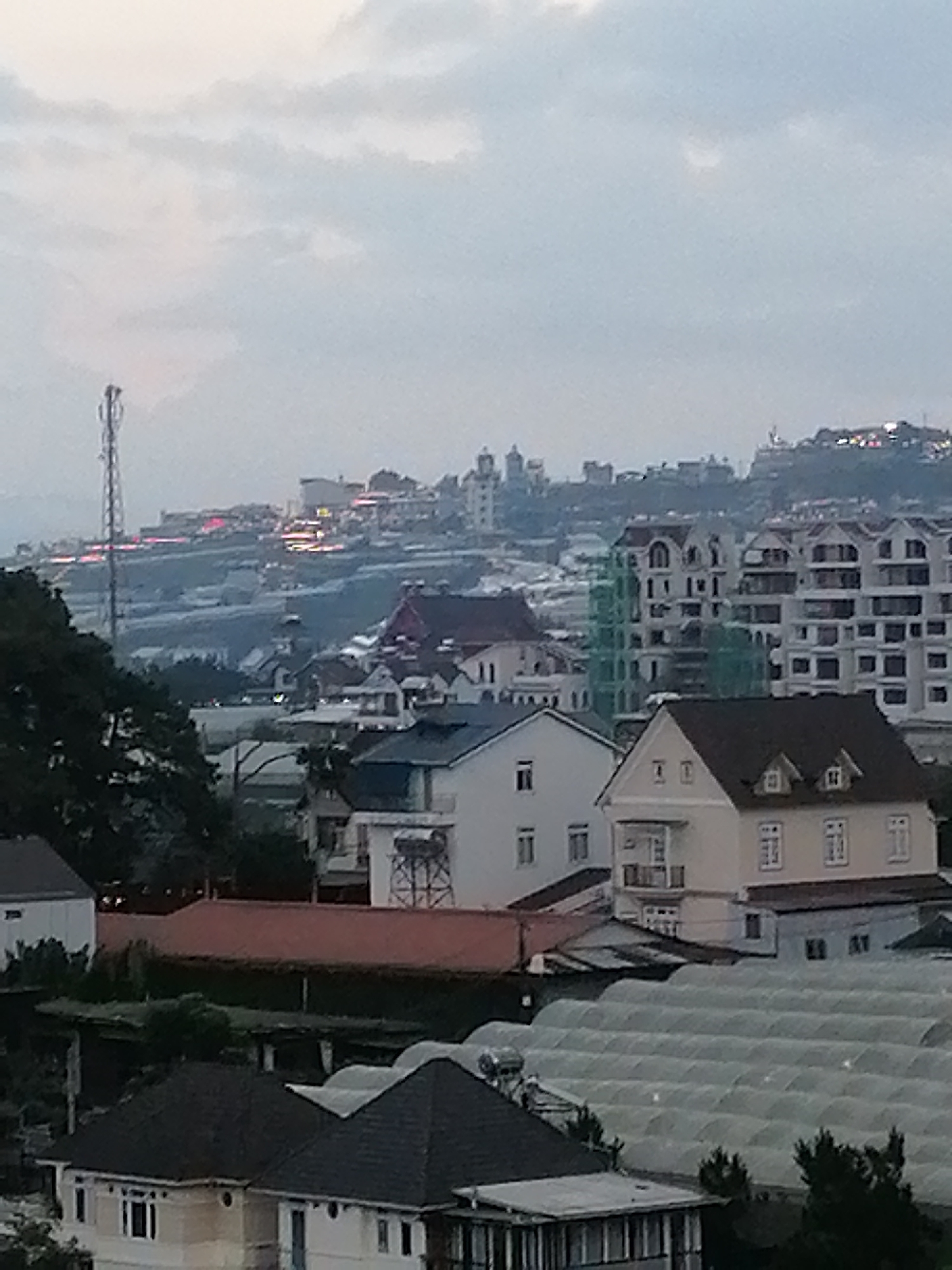 The image shows a panoramic view of a city or town on a hillside under a cloudy sky. In the foreground, there are several houses with dark roofs, some with white or light-colored walls. Further up the hill, more buildings are visible, including apartment complexes with many windows and some with balconies. A telecommunication tower stands tall on the left side of the image. In the distance, more buildings are scattered across the hillside, and some lights are visible, suggesting it might be dusk or dawn. The overall scene depicts a residential area with a mix of architectural styles. The hint images suggest that this location is Xuân Trường - Đà Lạt, Vietnam, known for its hilly terrain and European-inspired architecture.
