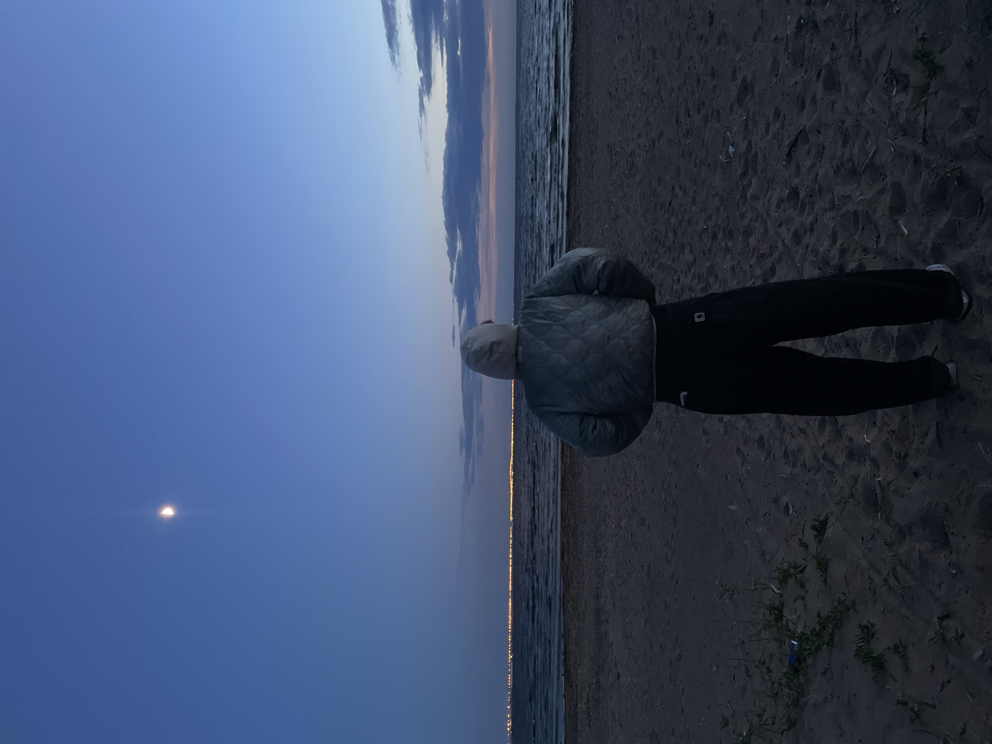 The image shows a person standing on a beach in Sestroretsk, Russia, at dusk or twilight. The individual is wearing dark pants and a light-colored, quilted jacket. The sky transitions from a deep blue to a lighter blue with some clouds on the horizon. The sea is visible with a line of lights across the water, possibly from a distant city. A small crescent moon is visible in the sky. The beach appears sandy. Some vegetation can be seen near the person’s feet.