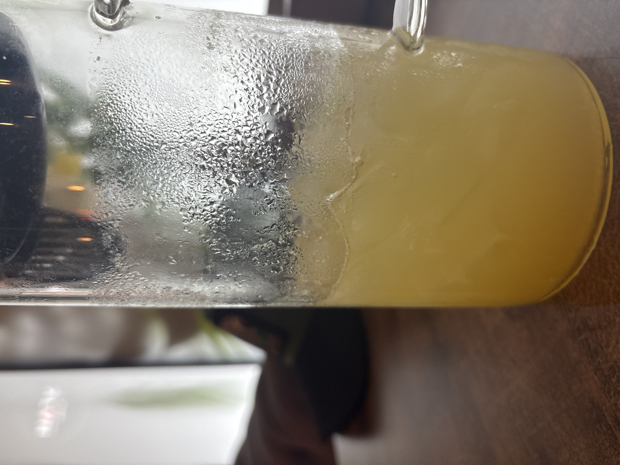 A close-up, eye-level shot shows a glass pitcher filled with a light yellow liquid and ice. The exterior of the pitcher is covered in condensation, obscuring the view of the background. The pitcher is placed on a brown, wooden surface. To the left, a dark object, possibly a computer mouse, and a blurred hand are visible, suggesting the pitcher is on a desk. The indistinct background shows a light-colored screen and hints of greenery, possibly indicating an indoor setting like an office or cafe. The image composition is tight on the pitcher, with no clear indication of the city of Chaikovsky, Russia.