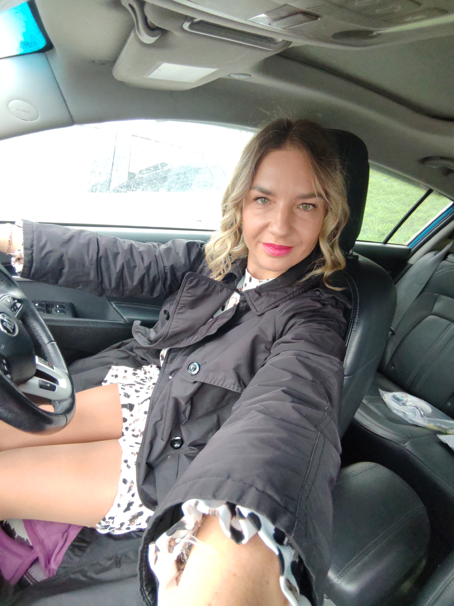 A woman is sitting in the driver's seat of a car. She is wearing a black jacket over a white and black patterned dress. She is holding the steering wheel. The interior of the car is visible, including the dashboard, seats, and windows. The outside is visible through the windows. The location is not discernible from this image.