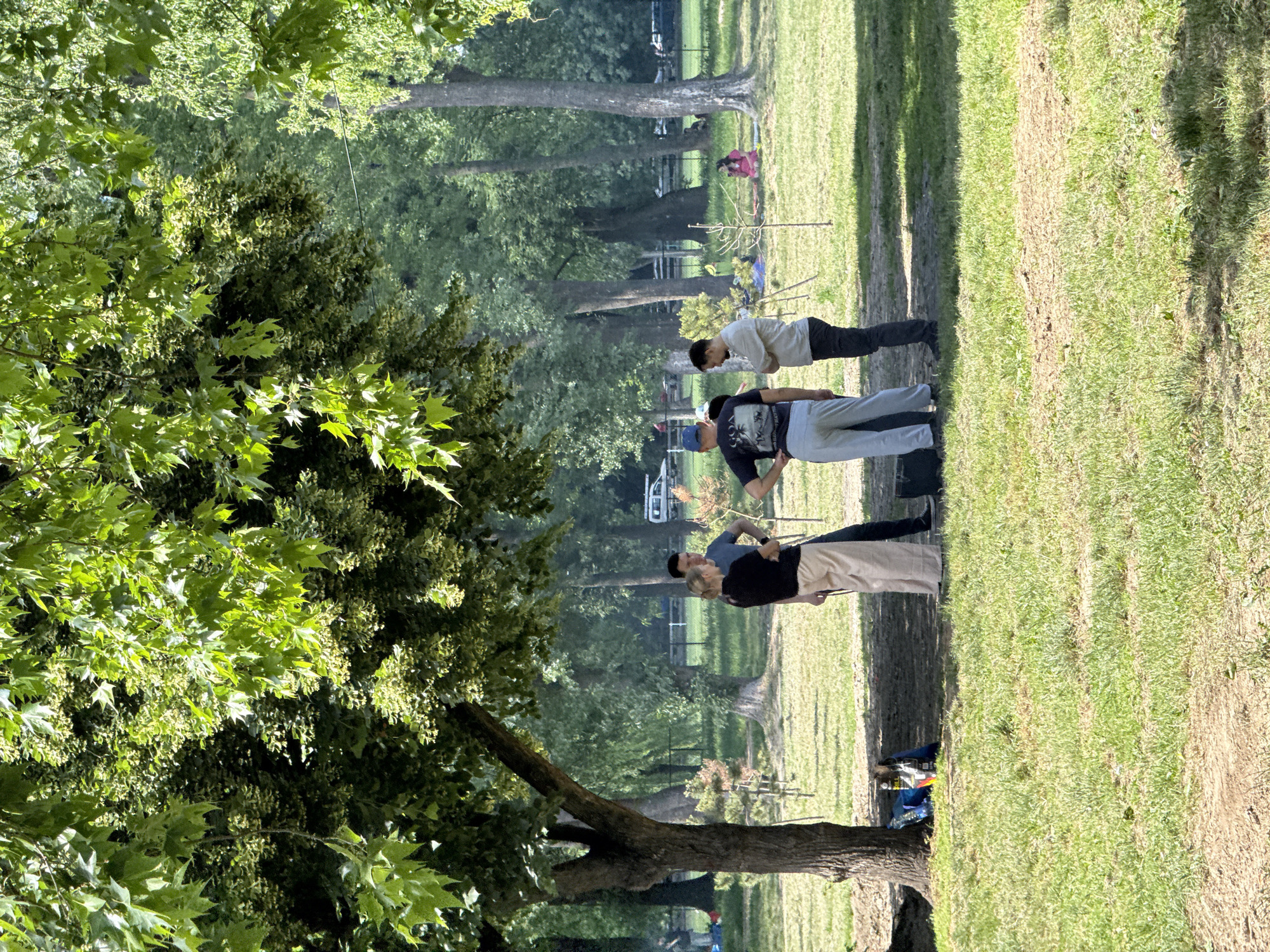 The scene appears to be set in a park-like area with grass and trees, possibly in Cukarica Urban Municipality, Serbia. Four people are standing in a group, seemingly conversing or observing something. One person wears a blue cap and dark shirt, while another sports a white shirt. The background shows more trees, and a woman seated with a pink jacket in the distance. A tree occupies a significant portion of the left side of the frame.