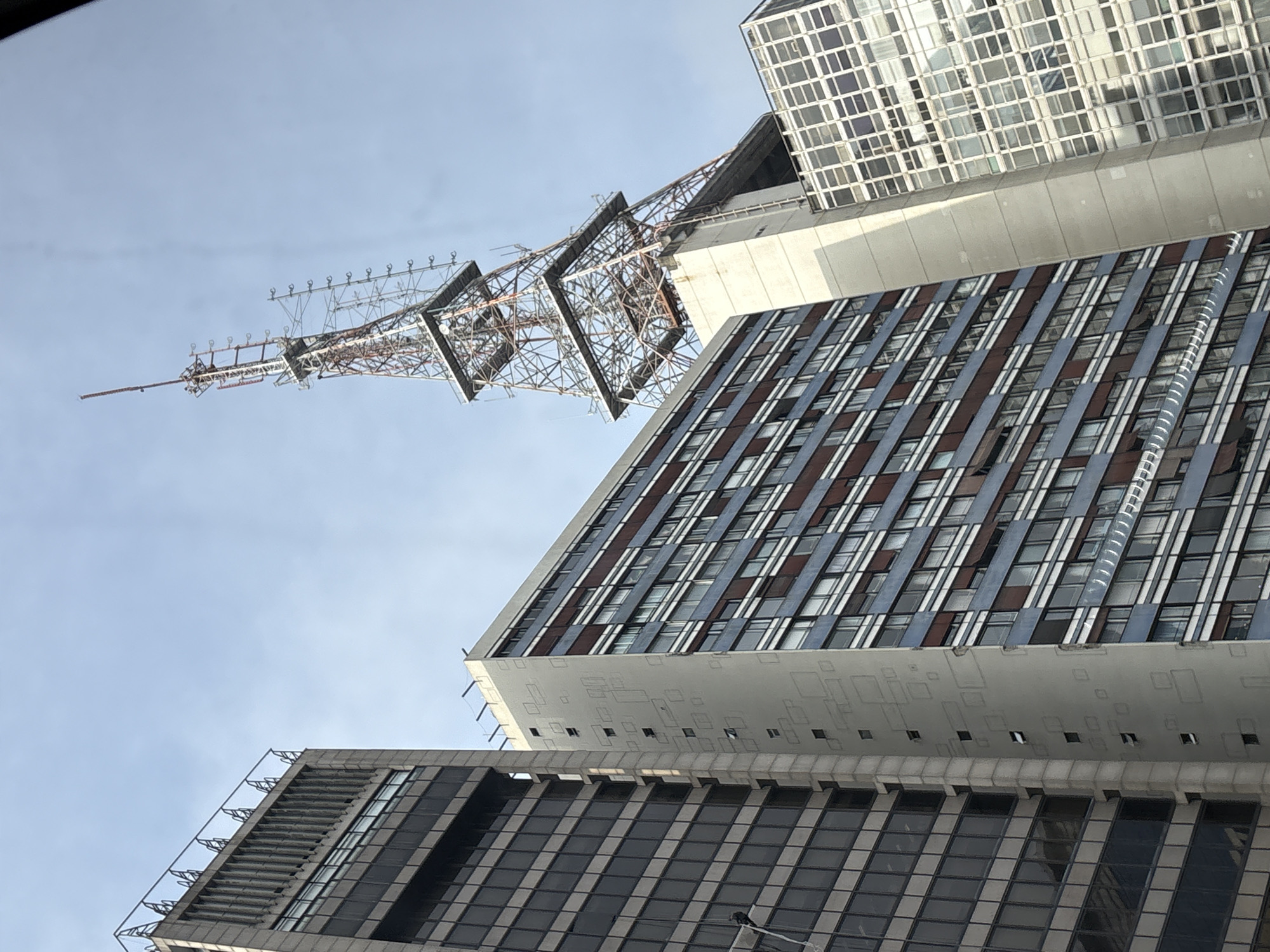 This is a low-angle, full-frame shot of a cluster of modern skyscrapers and a tall broadcast antenna in São Paulo, Brazil, under a clear, pale blue sky. In the foreground on the left is a dark glass-and-steel skyscraper with horizontal bands of windows and a vertical grid pattern. To its right is a lighter-colored building with a patterned facade, featuring irregular rectangular shapes and small, open windows on a light gray section. To the right of that is a blue and white building with rows of windows. Dominating the background is a large, lattice-style broadcast tower with multiple antenna arrays. The scene appears to be a business or downtown district. There are no people or animals visible in the image.