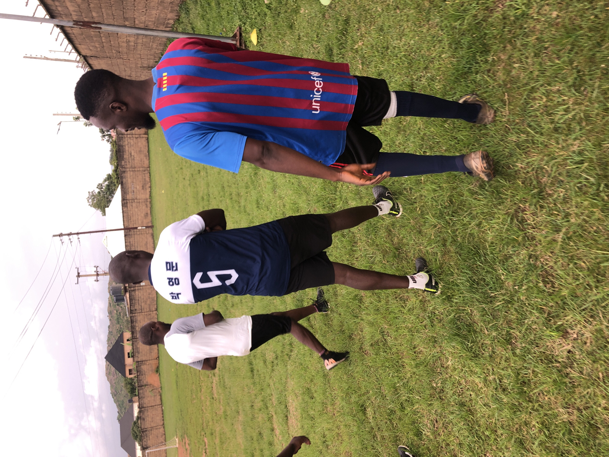 The image shows three young Black men wearing athletic attire on a grassy field, likely a soccer field, in Jos, Nigeria. The man in the foreground on the right is wearing a blue and red striped soccer jersey with "unicef" written in white text on the back, and black shorts with blue socks. He is looking to his left and smiling. The man in the middle is wearing a blue and white soccer jersey with Korean lettering and the number 5 in white on the back, and black shorts. He is walking away from the camera, looking to his left. The third man, on the left, is wearing a white and gray t-shirt and black shorts, and is also walking away from the camera, looking to his left. In the background, there is a brick wall, a soccer goalpost, some buildings, and hills. The sky is overcast. The field appears to be a community or school soccer field in a semi-urban area of Jos.