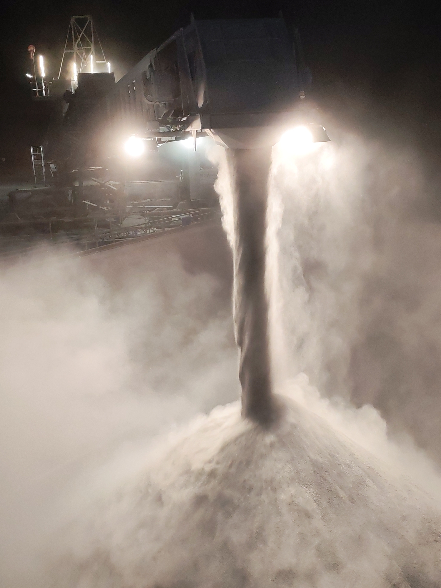 At night in Shcheleiki, Russia, a large industrial machine is dispensing a white substance, likely salt or similar material for winter road maintenance, onto a growing pile. The machine has bright lights illuminating the stream and the resulting mound. Mist or dust surrounds the activity, obscuring some details of the machinery and the surrounding environment. A metal structure with ladders and platforms is visible to the left of the machine.