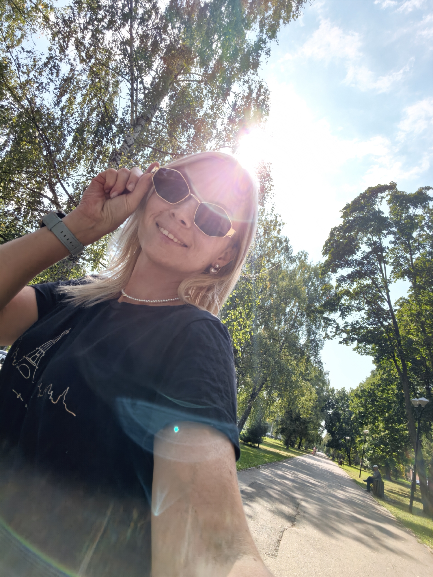 A woman with blonde hair, wearing sunglasses, a black t-shirt with a graphic design, and a pearl necklace, takes a selfie outdoors. She is holding her sunglasses with her right hand, and her left arm is visible with a grey smartwatch. The background features a paved path lined with trees and a clear sky with some clouds, suggesting a park or a street. The sun is shining brightly, creating lens flares. In the distance, a person is walking on the path, and another person is sitting on a bench to the right. The image is taken from a low-angle perspective. The presence of a park suggests the location could be a recreational area within a city. Based on the context provided, this scene is located in the city of Sterlitamak, Russia.