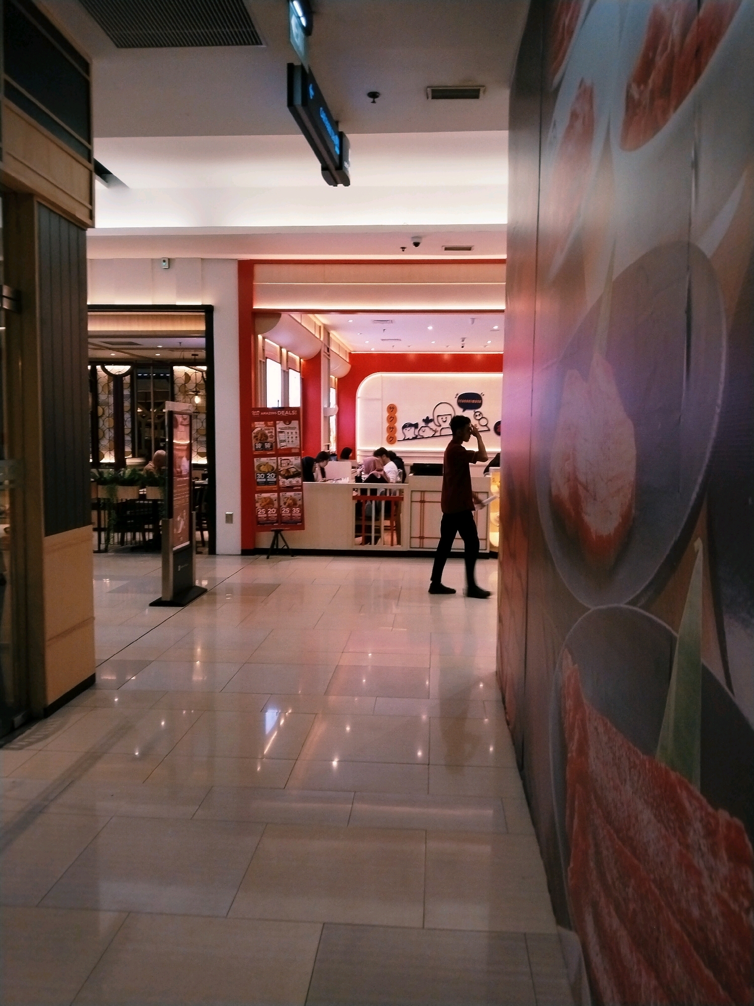 This image depicts a wide shot of a clean, well-lit interior walkway with large, reflective floor tiles. The walkway appears to be inside a commercial building, possibly a shopping mall or a food court, located in Surabaya, Indonesia. On the right side of the image, a large, colorful mural covers the wall, featuring abstract shapes and imagery resembling food items.
To the left, a portion of a building facade is visible, with darker wood accents and glass panels. Further down the walkway, on the left, is an entrance to a restaurant with a warm, inviting interior featuring decorative lighting. In the mid-ground, on the right side of the walkway, is a food establishment with a red and white color scheme. A menu board displaying prices and food images is prominently displayed outside. Several people are visible inside this establishment, seated at tables or queuing.
A lone man in a dark shirt and pants is walking through the walkway towards the right of the frame. He is seen from the side, with his hand near his face. The overall atmosphere is one of a modern, bustling commercial space.