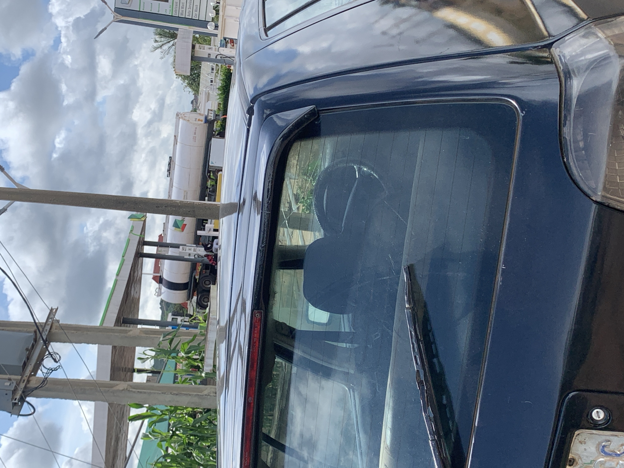 An outdoor medium shot shows the rear of a dark blue sedan parked at a gas station. The car is facing left and is angled slightly away from the camera. The car's rear window is visible, with a black headrest and part of a steering wheel inside. A windshield wiper is on the exterior of the rear window. Behind the car, a white tanker truck is parked under a green and white gas station canopy. The sky above is cloudy with patches of blue. Corn stalks are visible to the left of the gas station. The location is a gas station in an unidentified city.