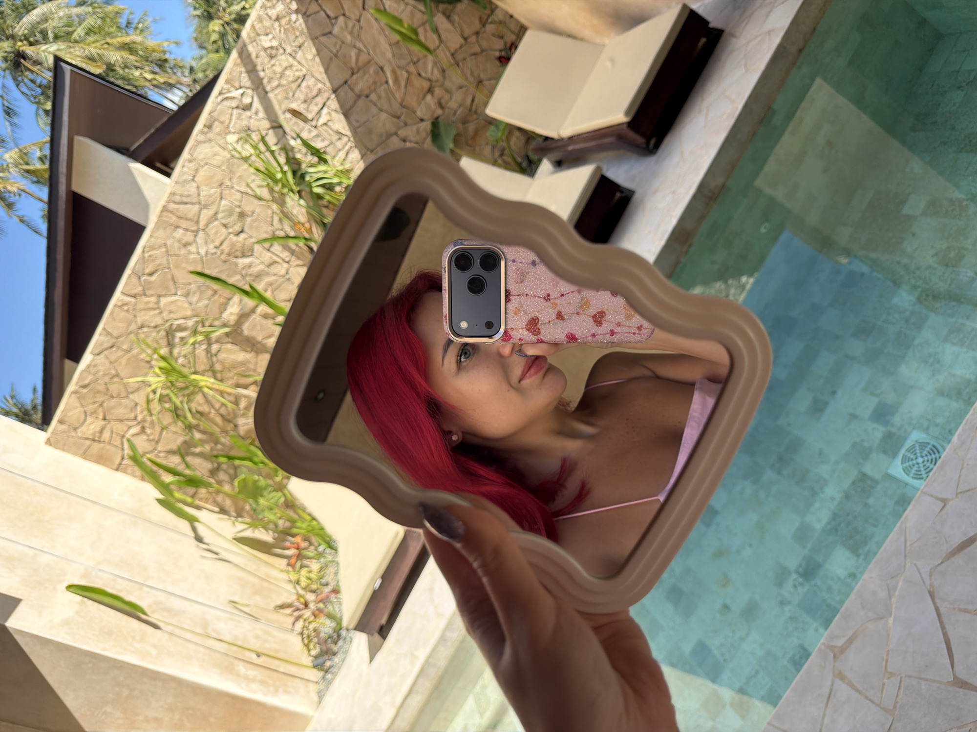 A woman with bright pink hair is taking a selfie in a wavy mirror. She is wearing a light pink camisole. The mirror reflects her face, upper body, and the phone she is holding, which has a pink heart-patterned case. The background visible in the mirror shows a swimming pool, lounge chairs, and a building with stone and cream-colored walls, accented by green plants. The image appears to be taken outdoors in a tropical setting, possibly Kuta, Indonesia, given the style of architecture and vegetation.