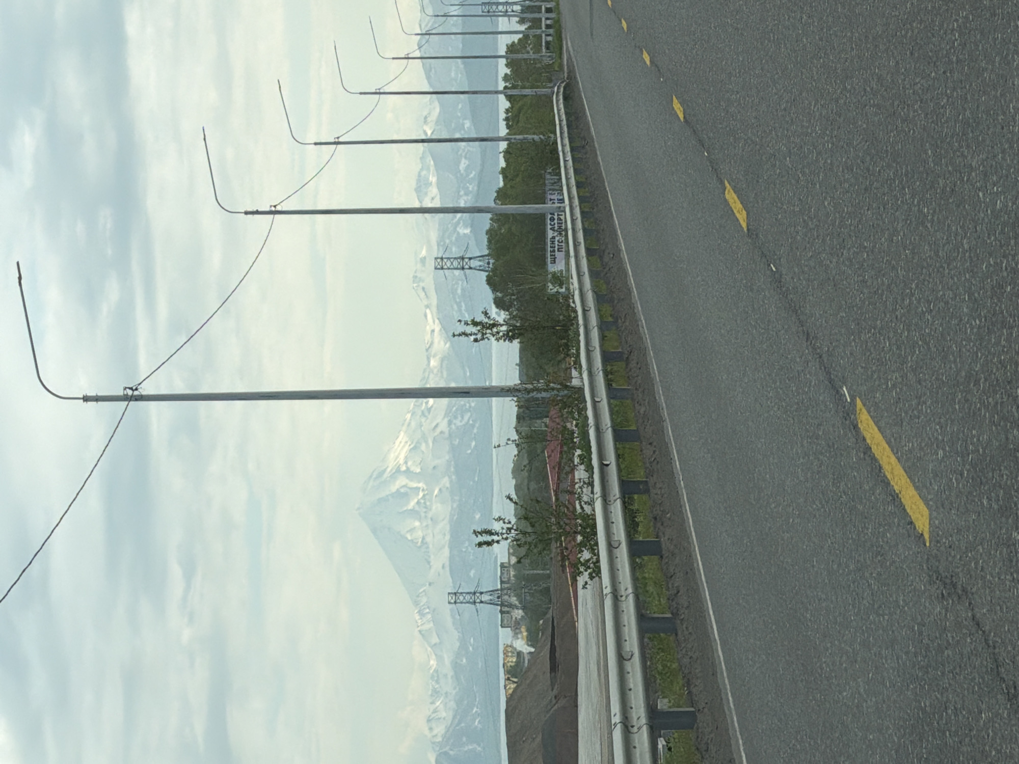 An eye-level, vertical medium shot captures a road leading towards a snow-capped mountain range under a cloudy sky. The road, surfaced with asphalt, features broken yellow lines as lane markers. On the left, a silver metal guardrail runs along the roadside, with a grassy embankment behind it. In the middle ground, several electrical transmission towers are visible, along with a body of water and lush green trees. A large, conical, snow-covered mountain dominates the background, with smaller, snow-capped peaks extending to its sides. The scene is likely located in or near the city of Svetly, Russia, given the prominent mountainous landscape.
