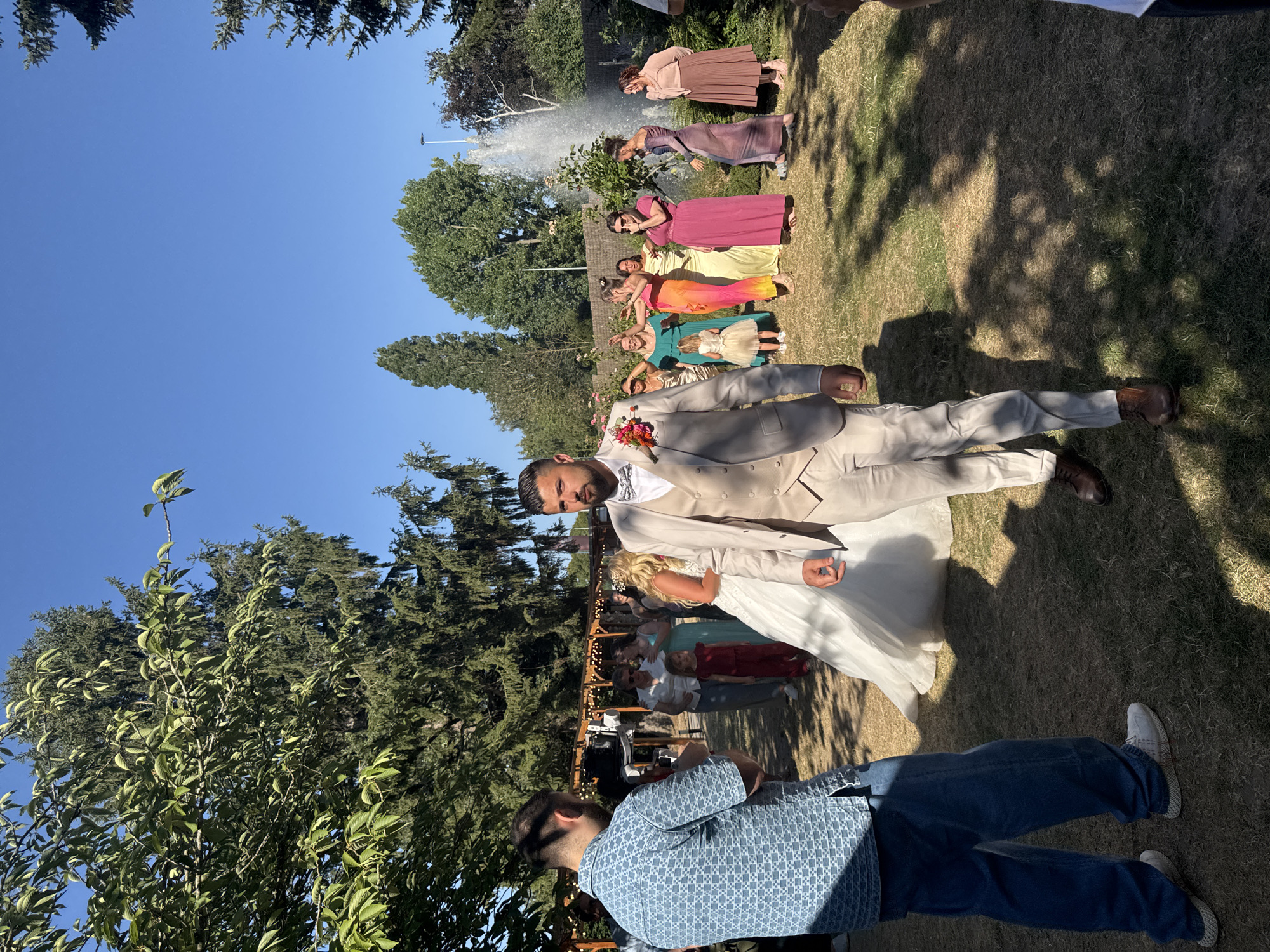 The image depicts an outdoor wedding ceremony in Hem, France. The scene is set on a grassy area surrounded by trees and features a fountain in the background. In the foreground, a groom in a light tan suit with a bow tie and a boutonniere is looking to his right. To his left, a man in a blue patterned shirt and jeans is holding a video camera. Several women in colorful dresses and a young girl in a red outfit are gathered in the midground, observing the ceremony. A bride in a white wedding dress is also visible, facing the groom. The atmosphere suggests a celebration with guests and a professional videographer capturing the event. The lighting indicates it is daytime, with shadows cast by the trees on the grass.