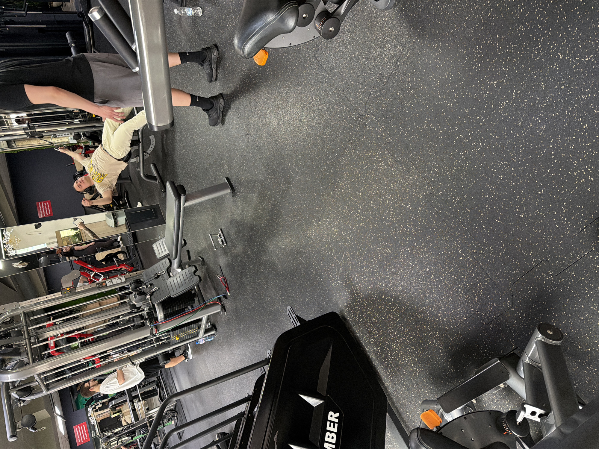 The image depicts the interior of a gym, likely located in a commercial area based on the presence of gym equipment and a mirrored wall indicating a designated workout space. The gym features various exercise machines including a cable machine, leg press, and other weight training apparatus. The floor is covered in dark, speckled rubber matting.
Several individuals are present, engaged in various activities. One person is seated on a cable machine, presumably performing an exercise. Another individual is standing nearby. In the mirrored wall, reflections show others using different gym equipment.
There are no identifying details that would suggest that this photo was taken in the city of Pleven, Bulgaria.