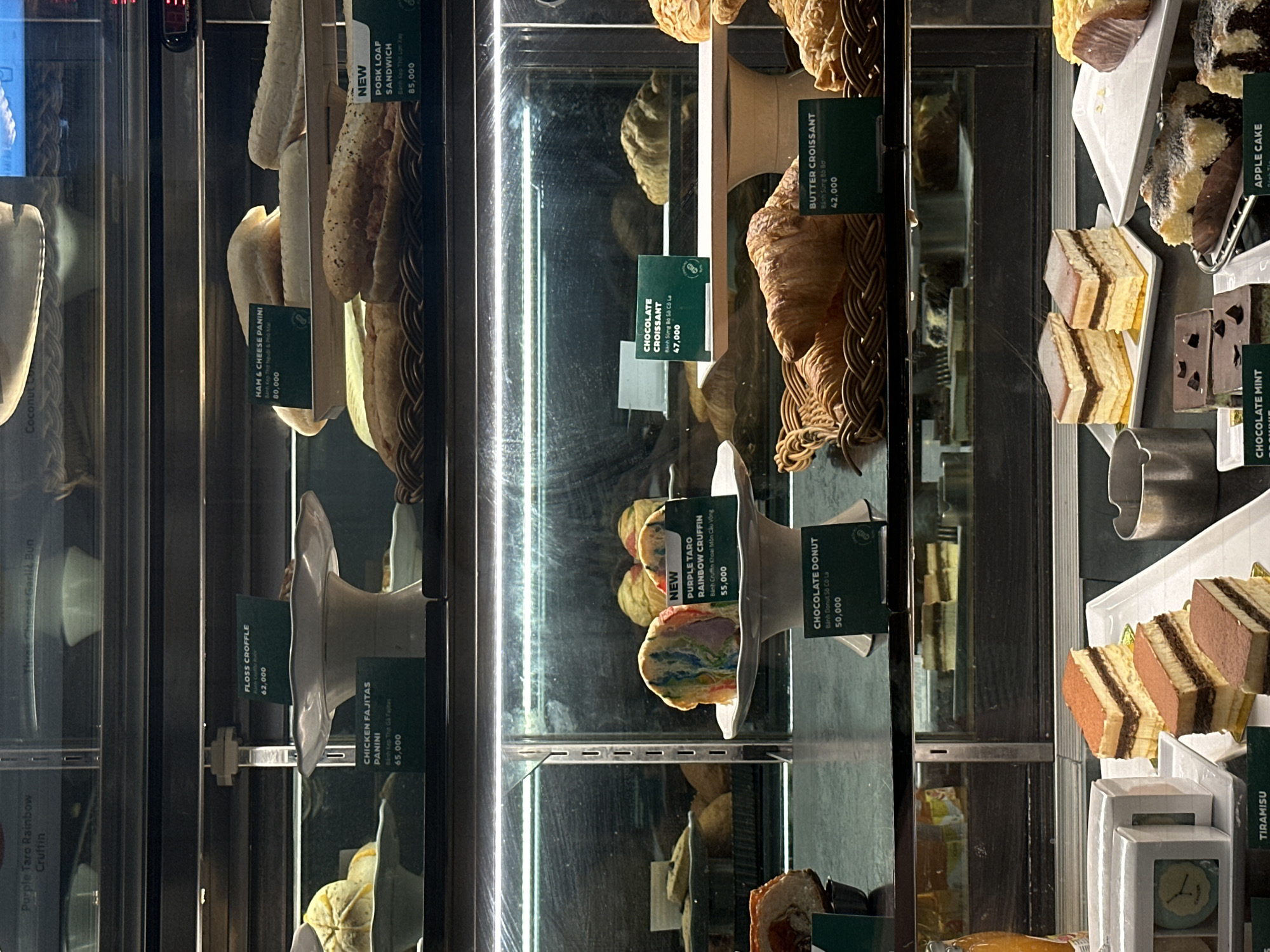 The image shows a display case filled with various pastries and sandwiches in a food establishment in Đà Nẵng, Vietnam. The display case is illuminated by overhead lights, creating a slightly shadowed effect.
In the top shelf, several baked goods are visible, including what appears to be a purple taro cruffin with rainbow swirls and a sign indicating "NEW PURPLE TARO RAINBOW CRUFFIN 55,000". To the right of this, there are ham and cheese paninis labeled "HAM & CHEESE PANINI 80,000". Further to the right, a "PORK LOAF SANDWICH 85,000" is displayed.
The middle shelf features more pastries. There is a "CHOCOLATE DONUT 50,000" and a "BUTTER CROISSANT 42,000". In the background of this shelf, several croissants are visible.
The bottom shelf displays several slices of layered cakes, including what appears to be tiramisu and other chocolate and cream layered desserts. A sign for "TIRAMISU" is partially visible at the bottom left. Also on this shelf are what look like chocolate mint brownies and apple cake.
The overall environment suggests a bakery or cafe, with the focus on a well-lit display of tempting food items. The signs are in English and Vietnamese, confirming the location in Vietnam.