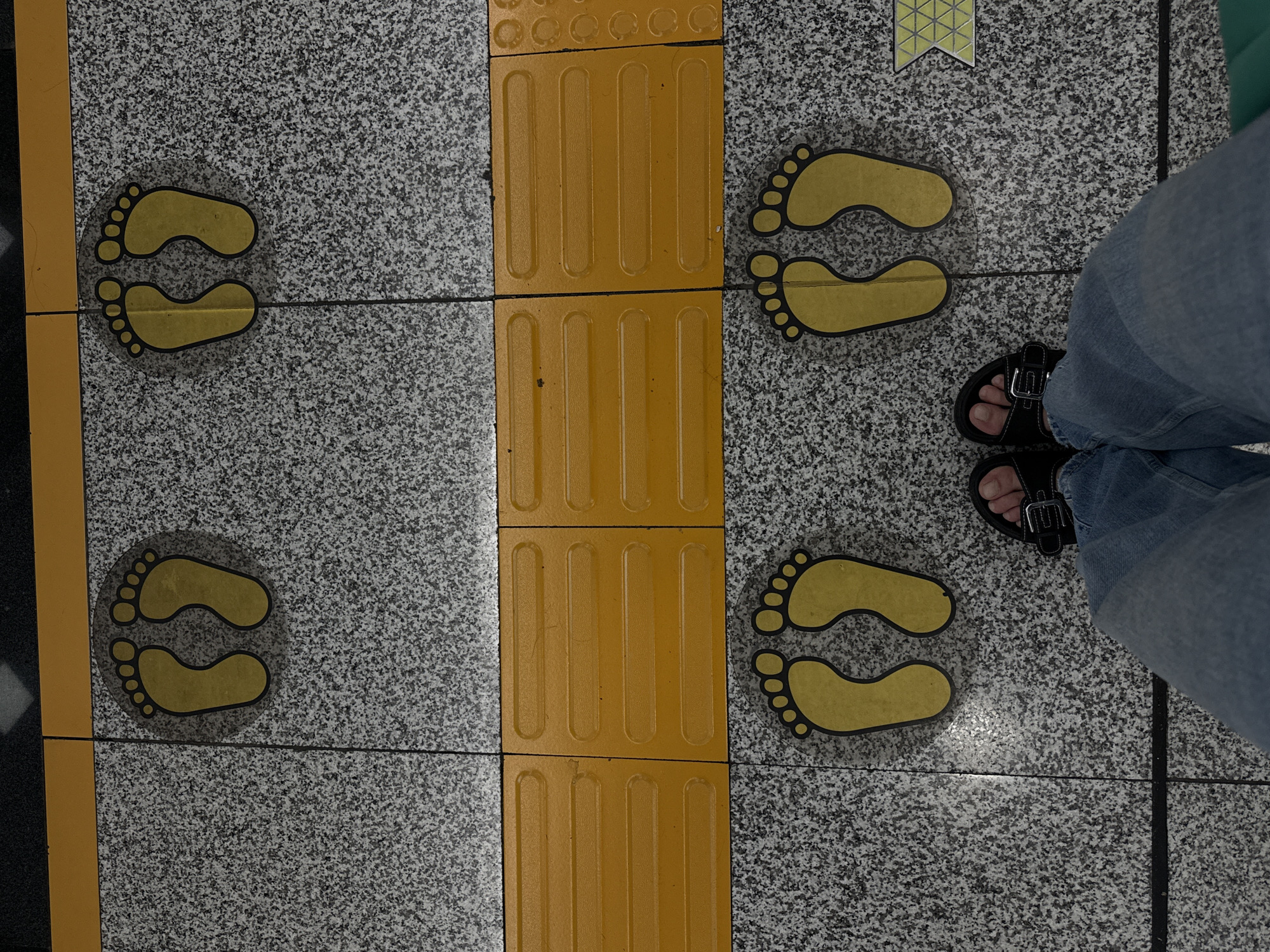 The image depicts a ground-level perspective of a public space, likely a subway station in Nanshan District, China, indicated by the presence of tactile paving. In the foreground, a person's legs, clad in blue jeans and black sandals, are visible, positioned between two sets of yellow footprint decals on a speckled gray tiled floor. These decals, indicating designated standing spots, are arranged in pairs, one set further back and slightly to the left, and another closer and centered. A bright yellow textured strip runs horizontally across the mid-ground, marking a boundary or safety zone. The background shows more of the speckled tiles and a dark line, further suggesting an urban transit environment.