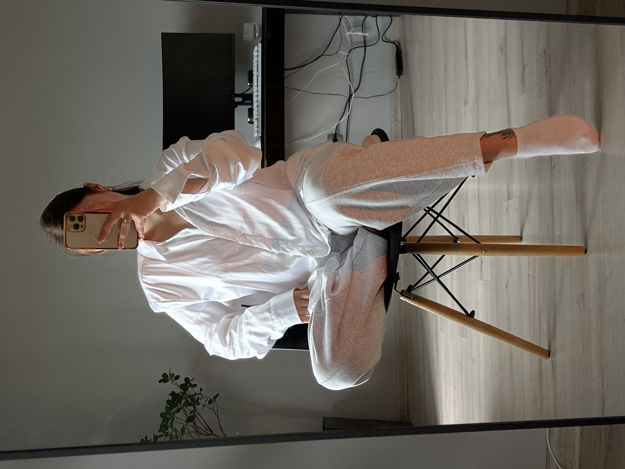 An indoor mirror selfie captures a person seated on a black chair with wooden legs, positioned in the city of Ternopil, Ukraine. The individual, whose face is obscured by a gold-cased smartphone held in their right hand, is wearing an oversized white button-down shirt and light grey sweatpants. Their left leg is crossed over the right, with the knee elevated. The right foot, clad in a light pink sock, is partially lifted, revealing a dark tattoo on the inner ankle. To the left of the reflection, a green leafy plant in a dark pot is visible. To the right, a black desk holds a computer monitor, a white keyboard, and a clear glass, with a tangle of power cables visible beneath. The room features light grey walls and a light wood-planked floor.