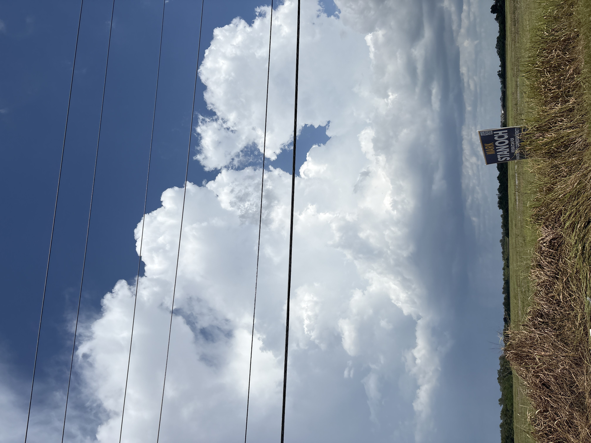 A political campaign sign for Mark Stanoch is visible in a field in Parrish, Florida, United States. The sign is blue and white with text that reads "MARK STANOCH" and "REPUBLICAN FOR COUNTY COMMISSIONER DISTRICT 1". The sign is situated in a grassy field with some dry grass in the foreground. In the background, there are trees and a cloudy blue sky with power lines running across the top of the image. The area appears to be rural.
