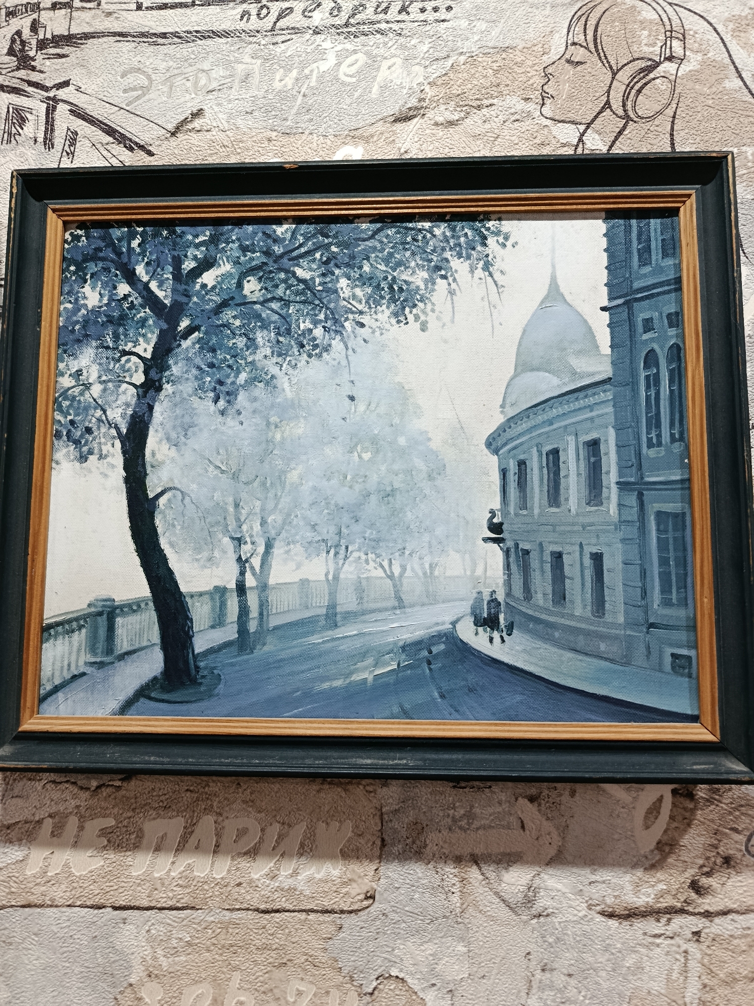 The image shows a framed painting depicting a street scene in what appears to be a city. The painting is rendered in muted blues and grays, suggesting a cool or possibly overcast day. On the left side of the painting, a large, dark tree with sparse foliage dominates the foreground. A curved stone wall with decorative elements runs alongside a pathway. In the background, several other trees are visible, creating a sense of depth. The right side of the painting features a light blue building with multiple windows and a domed structure behind it. Two figures are walking on the street in front of the building. The painting is framed with a dark outer frame and a lighter inner frame. The painting hangs on a textured wall. The wall has a pattern resembling wallpaper with some text and drawings, and the lower portion of the wall is textured like brickwork. The overall impression of the painting is one of quiet contemplation within an urban setting. The location in the city of Apatity, Russia is not discernible from the image itself.