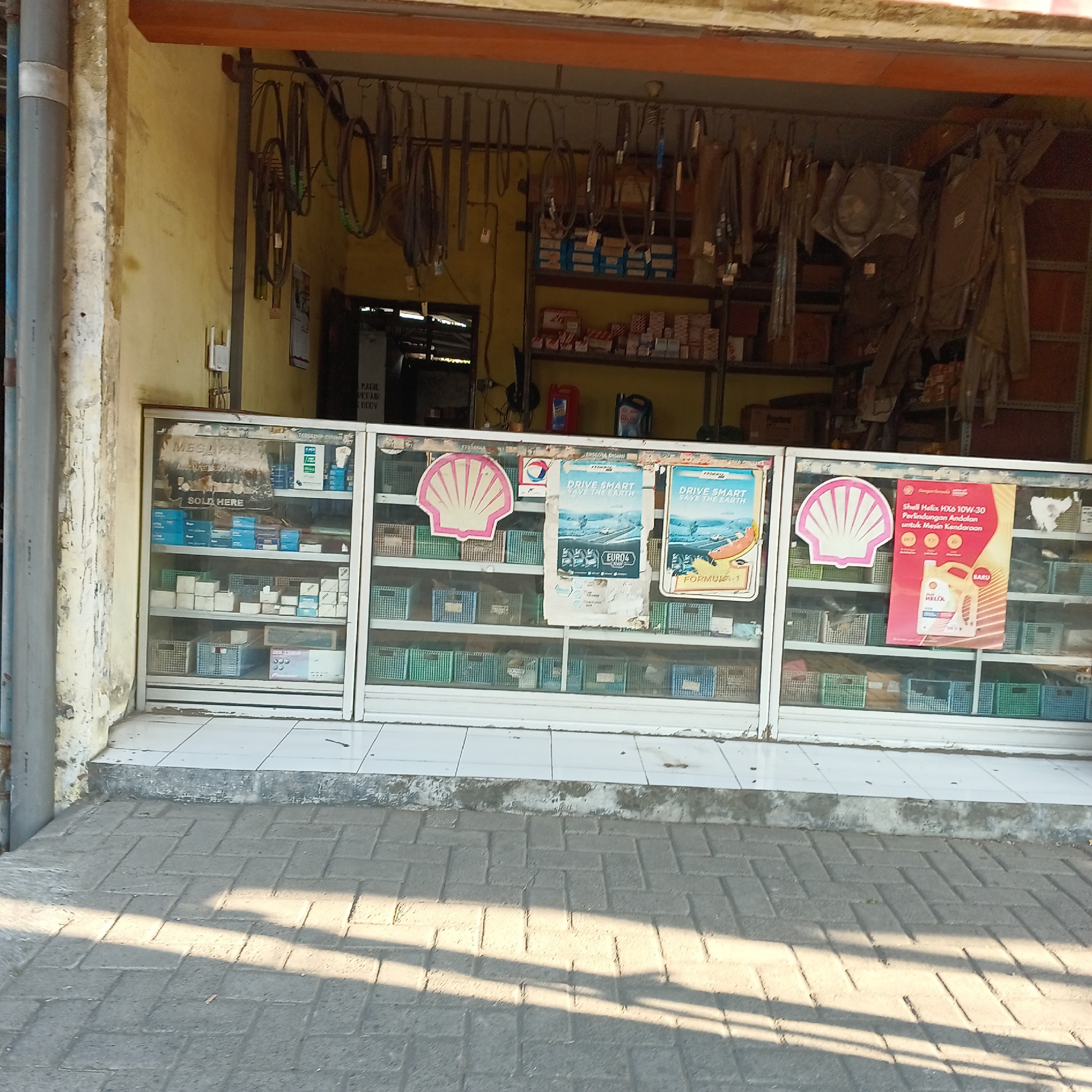 The image shows a storefront in Wedoro, Indonesia, displaying automotive parts and supplies. The exterior is weathered, with a pale yellow wall and a faded orange awning. In front of the store, a sidewalk paved with interlocking bricks is visible, with a shadow cast from the awning. The storefront itself features a glass display case filled with various items, including boxes, baskets, and posters. One poster advertises Shell Helix engine oil. Above the display case, belts and other automotive parts hang from a rack. The overall scene suggests a small, local auto parts shop.