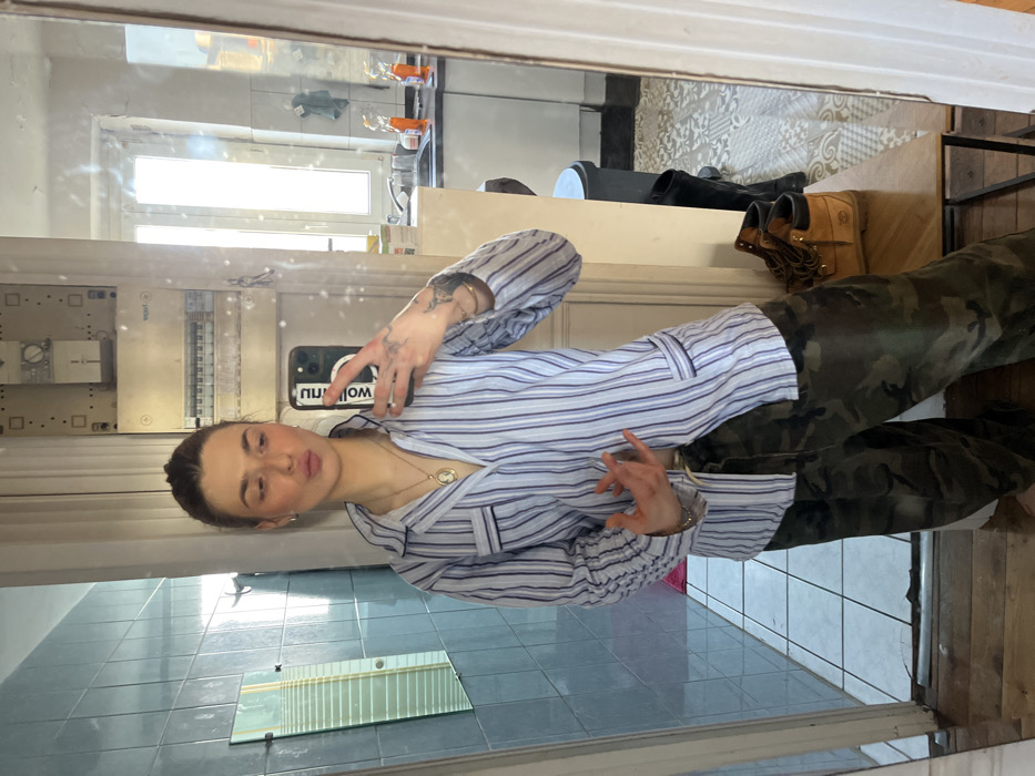 A young woman is captured in a mirror selfie, standing upright and slightly angled. She has dark hair pulled back, exposing her face which features light makeup and a "kiss" pose with pursed lips. She has freckles visible across her nose and cheeks.
She is wearing a light blue and white striped long-sleeved button-up shirt, which appears to be styled like pajama top, complete with a collar and pockets on the chest. A gold chain necklace with a cameo-style pendant hangs around her neck, and she also wears earrings, rings on her fingers, and a bracelet on her left wrist. Tattoos are visible on her left arm and hand.
Her lower body is clad in camouflage-patterned pants.
In her right hand, she holds a black smartphone, possibly an iPhone, with its camera lens module visible. The phone's screen or case reflects some reversed text, primarily "WOLF" and "NU", suggesting "WOLF NUDIE" or a similar brand/phrase. Her left hand is raised slightly, with fingers gently bent.
The setting is reflected in the mirror, revealing an indoor space. Behind the woman's reflected image:
* To the upper left, a bright window is visible, letting in strong daylight and showing a blurred view of what might be an urban environment or distant buildings outside.
* Below the window, there's an electrical panel or fuse box mounted on a light-colored wall.
* Further down, a light blue tiled wall is visible, and at the bottom, a rectangular mirror reflecting the deeper part of the room.
To the right side of the mirror's reflection, behind the camera/viewer, several objects are placed on a surface. These include a pair of light brown, work-style boots (resembling Timberlands) and other darker shoes. A section of patterned wallpaper or textured wall can also be seen. The floor beneath the shoes appears to be made of wood.
The overall lighting suggests it is daytime. The image conveys a casual, self-portrait moment.