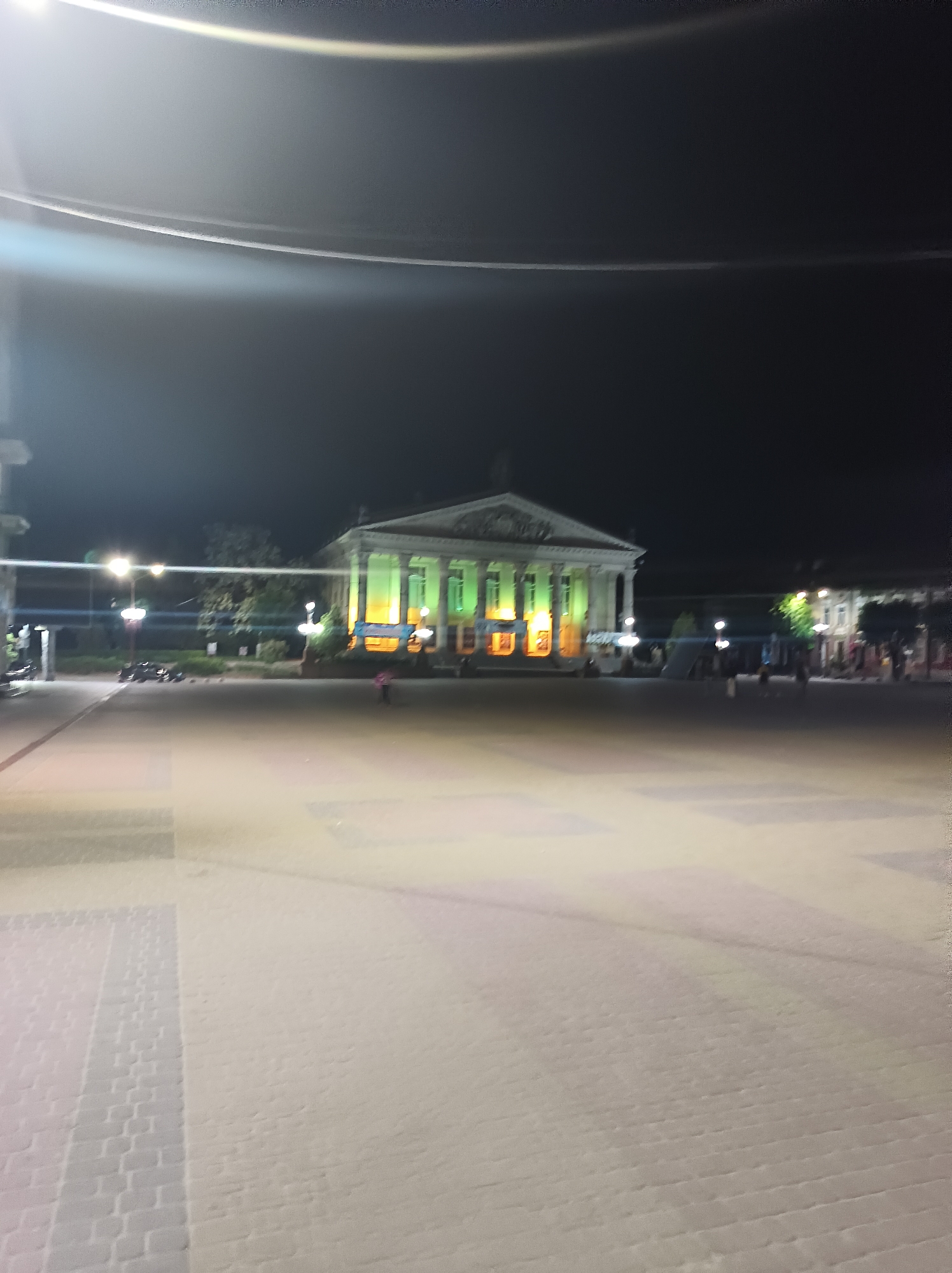The image shows Teatralnyi Square in Ternopil, Ukraine at night. The Ternopil Academic Drama Theatre, illuminated with green and orange lights, is the prominent structure. In the foreground, there is a large paved square. Several figures are seen in the distance. Streetlights provide additional illumination. The sky is dark.