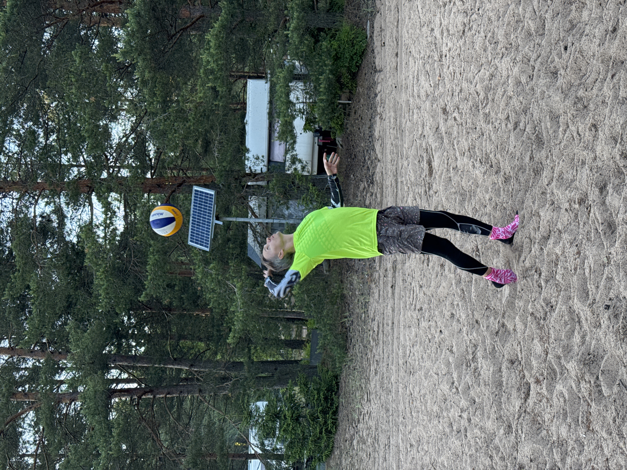 A person wearing a bright green shirt and black leggings with pink socks is playing volleyball on a sandy court. They are in the process of hitting or receiving the ball, which is in the air above them. A volleyball net is not visible. In the background, there are pine trees and a small white building, possibly a trailer or shed. A solar panel is mounted on a pole near the volleyball court. The scene appears to be outdoors, likely in a natural setting given the trees and sand. The lighting suggests it could be daytime, with overcast or filtered sunlight. The person's posture indicates they are actively engaged in the sport, with a focused expression. The overall atmosphere seems casual and recreational.