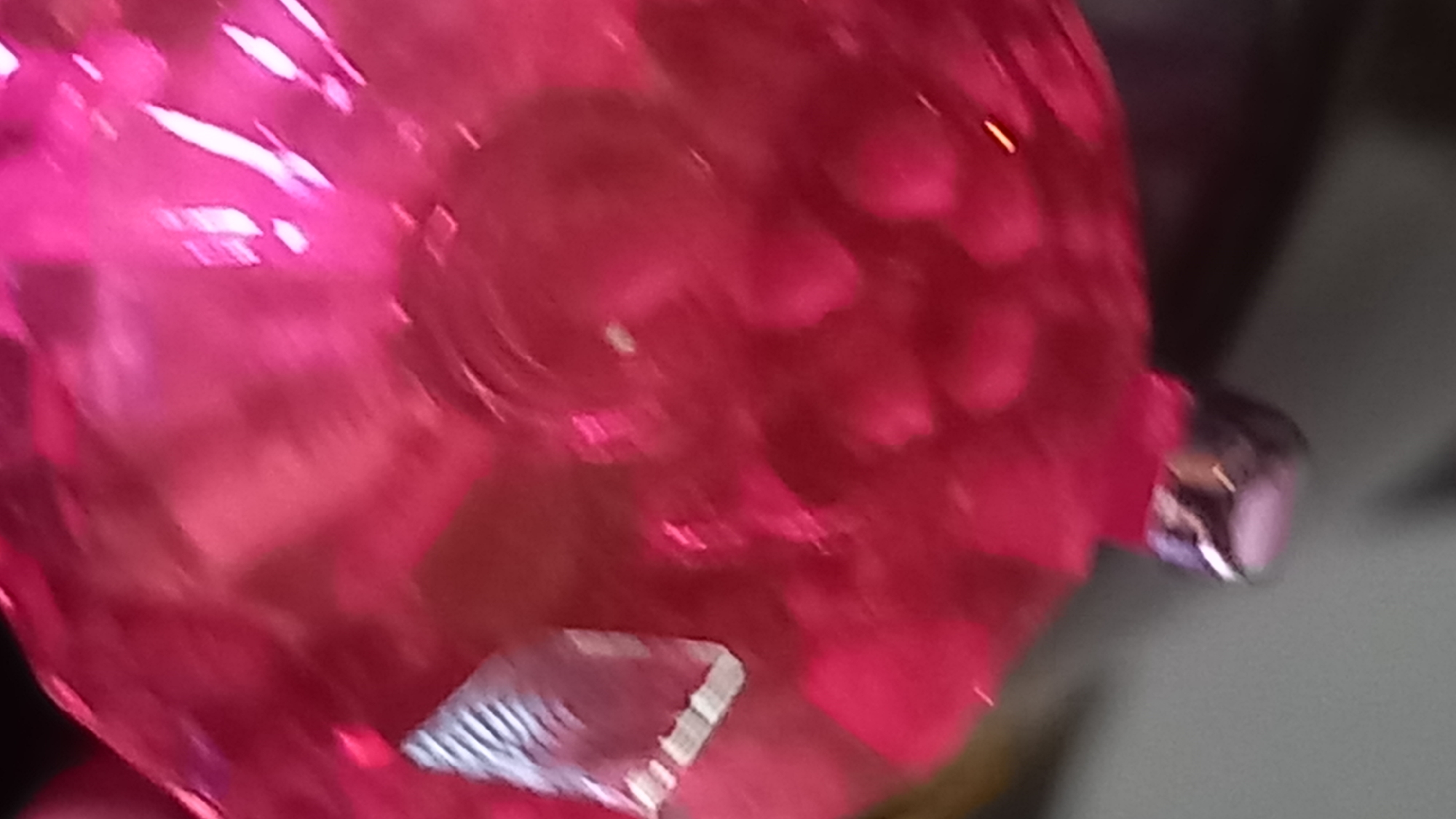 This is a close-up, out-of-focus photograph of a shiny, translucent pink object, possibly a decorative ornament or a piece of fabric. The object appears to have a textured surface with some raised bumps. A small, metallic, cylindrical object with a silver and purple hue hangs from the bottom of the pink object. The background is dark and blurred, offering no discernible details about the location. It is impossible to determine the specific location in the city of Kos, Greece from this image.