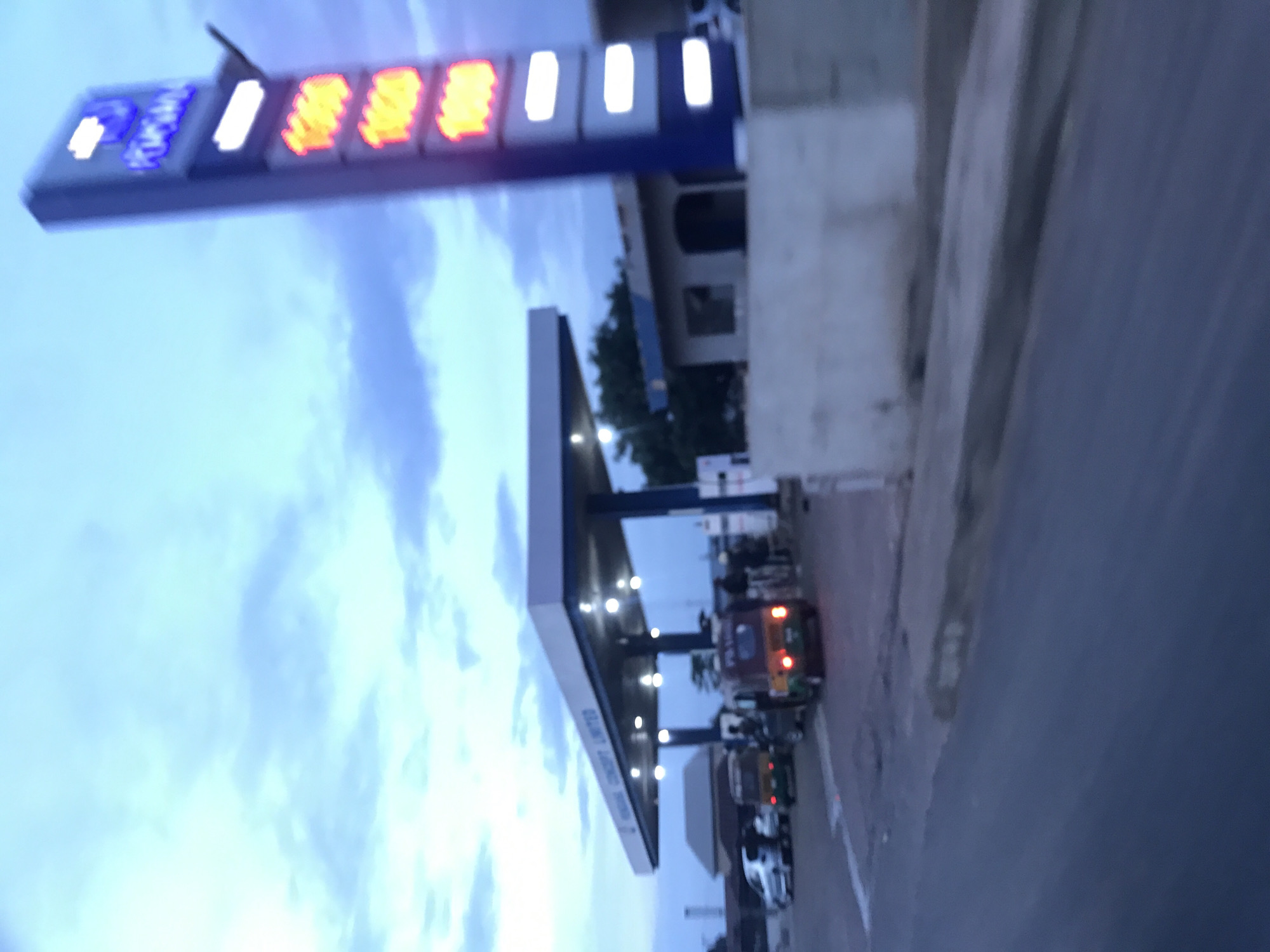 The image depicts a gas station identified as "KONGA CONCEPT LIMITED" in Rafin Par, Nigeria, under low light conditions consistent with dusk or dawn. A prominent vertical sign on the right side displays the name "KONGA" at the top, followed by three illuminated orange digital displays, each showing "1660," presumably indicating fuel prices. Below these, two blank white illuminated rectangular segments are present.
The gas station features a white canopy supported by blue pillars, under which multiple lights are illuminated. Several vehicles are present: two tricycles (auto-rickshaws) with green and brown/orange coloring are positioned under the canopy, facing away from the viewer. A silver sedan is parked further back to the left, and another dark-colored vehicle is partially visible. Multiple indistinct human figures are discernible under the canopy, interacting within the station area. The foreground shows a paved road surface with a visible white line and a low, light-colored concrete barrier separating the road from the station. The sky above is partially cloudy with shades of light blue and grey. A small, light-colored building with a dark roof is visible behind the price sign, to the right of the main canopy.