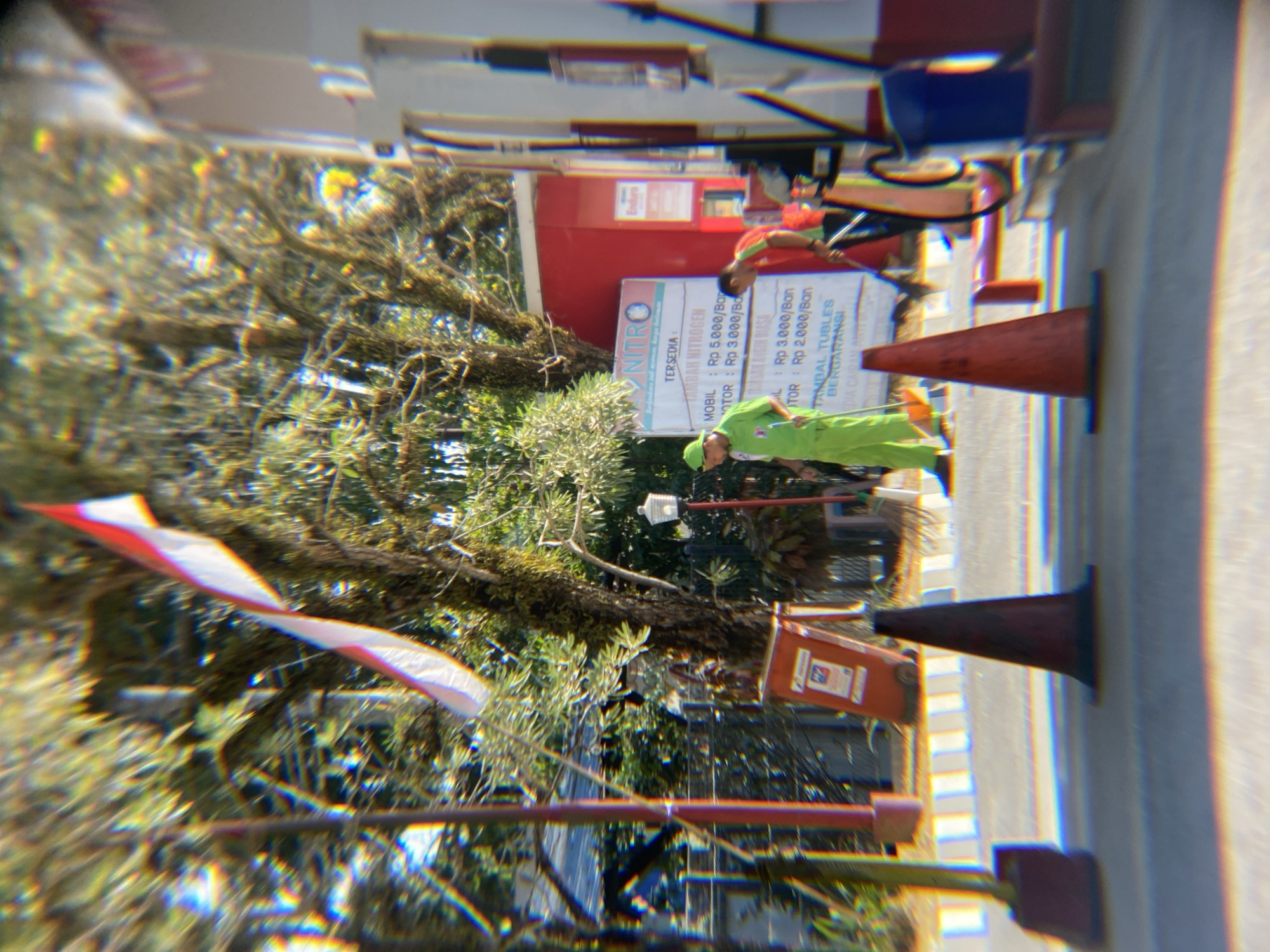 The image depicts an outdoor scene at what appears to be a gas station, possibly in Kendari, Indonesia. In the foreground, there are two orange traffic cones on the asphalt. A person wearing a bright green uniform and cap is sweeping the ground with a broom. Behind this person, another individual in a red and green uniform is also visible. A red A-frame sign is placed on the ground to the left. Further back, a tall flag pole with a red and white flag is visible, along with trees and buildings. The overall atmosphere suggests a sunny day.